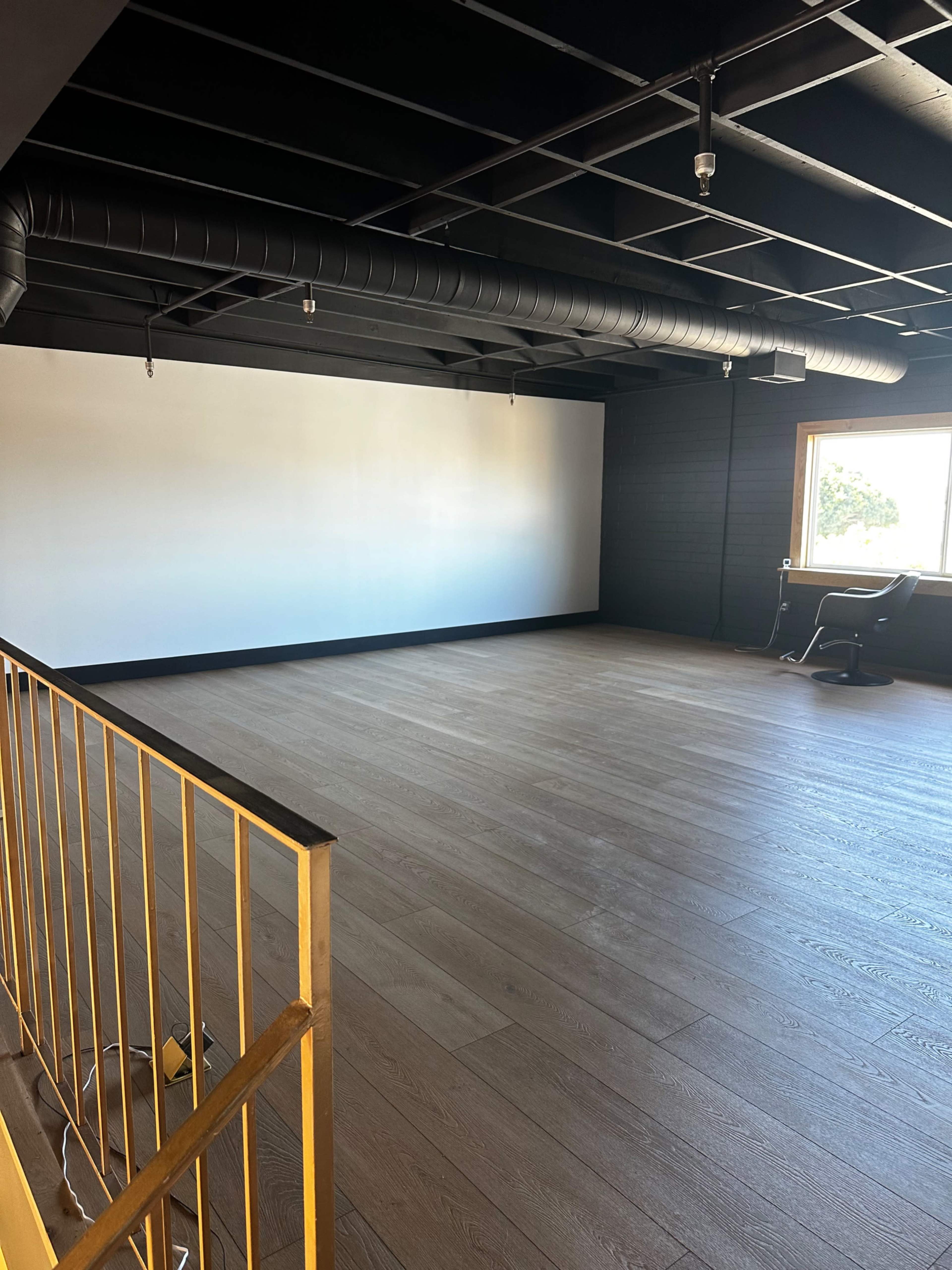 An empty office space features a large white wall, a wooden floor, and a single chair near a window.