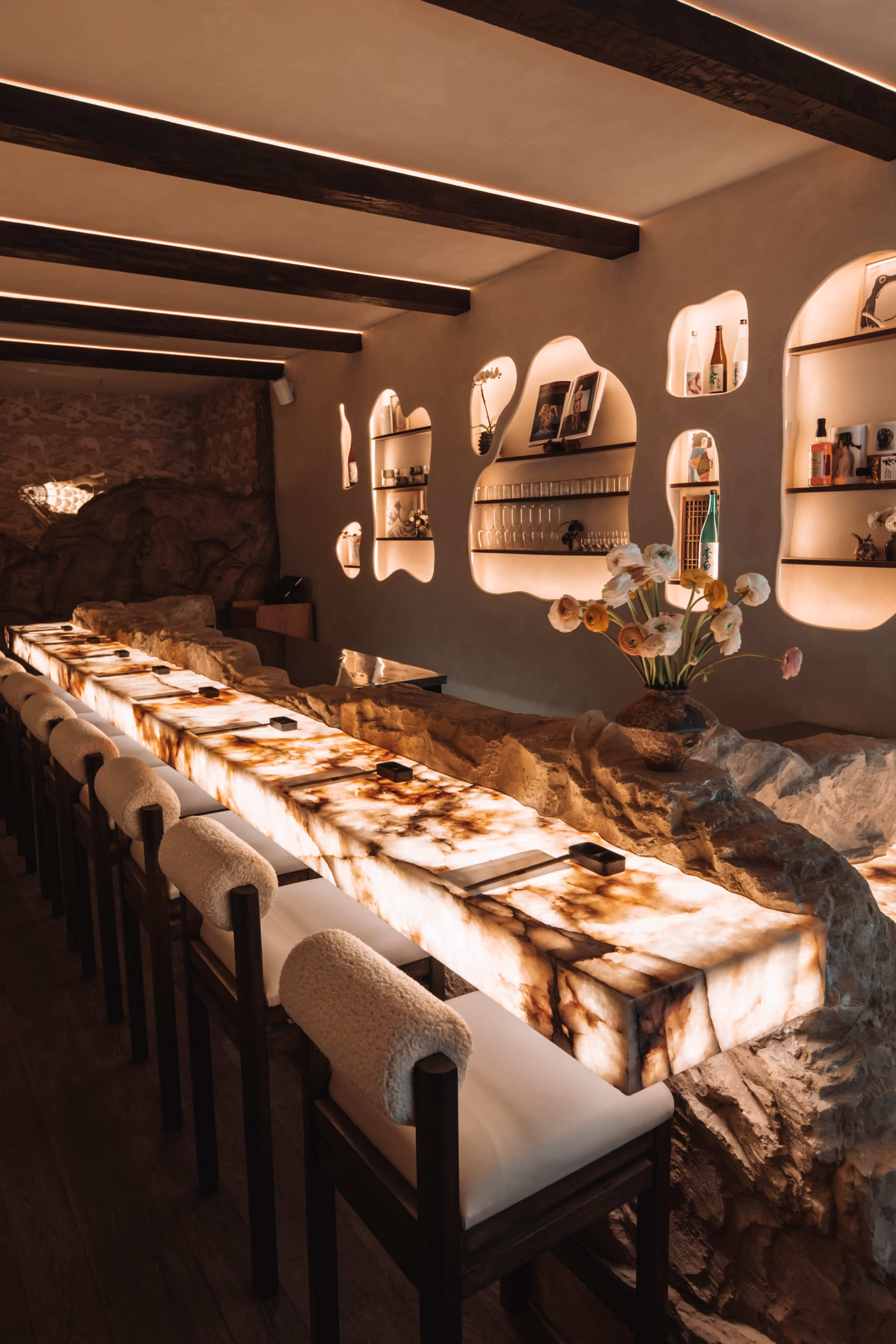 The image shows a modern restaurant interior with a long illuminated stone counter and shelves set into the walls featuring decorative items.