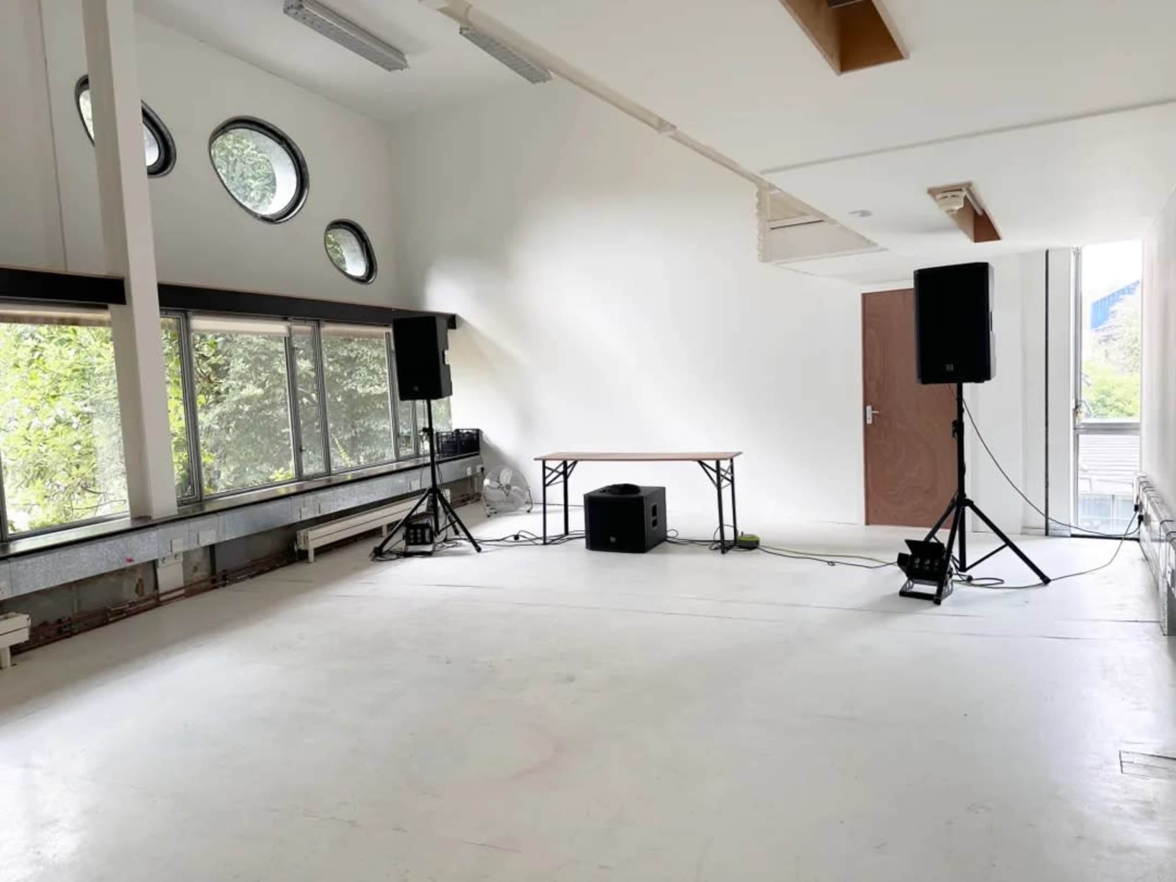 The image shows a minimalistic, brightly lit room with two speakers on stands, a small table in the center, and large windows along one wall.