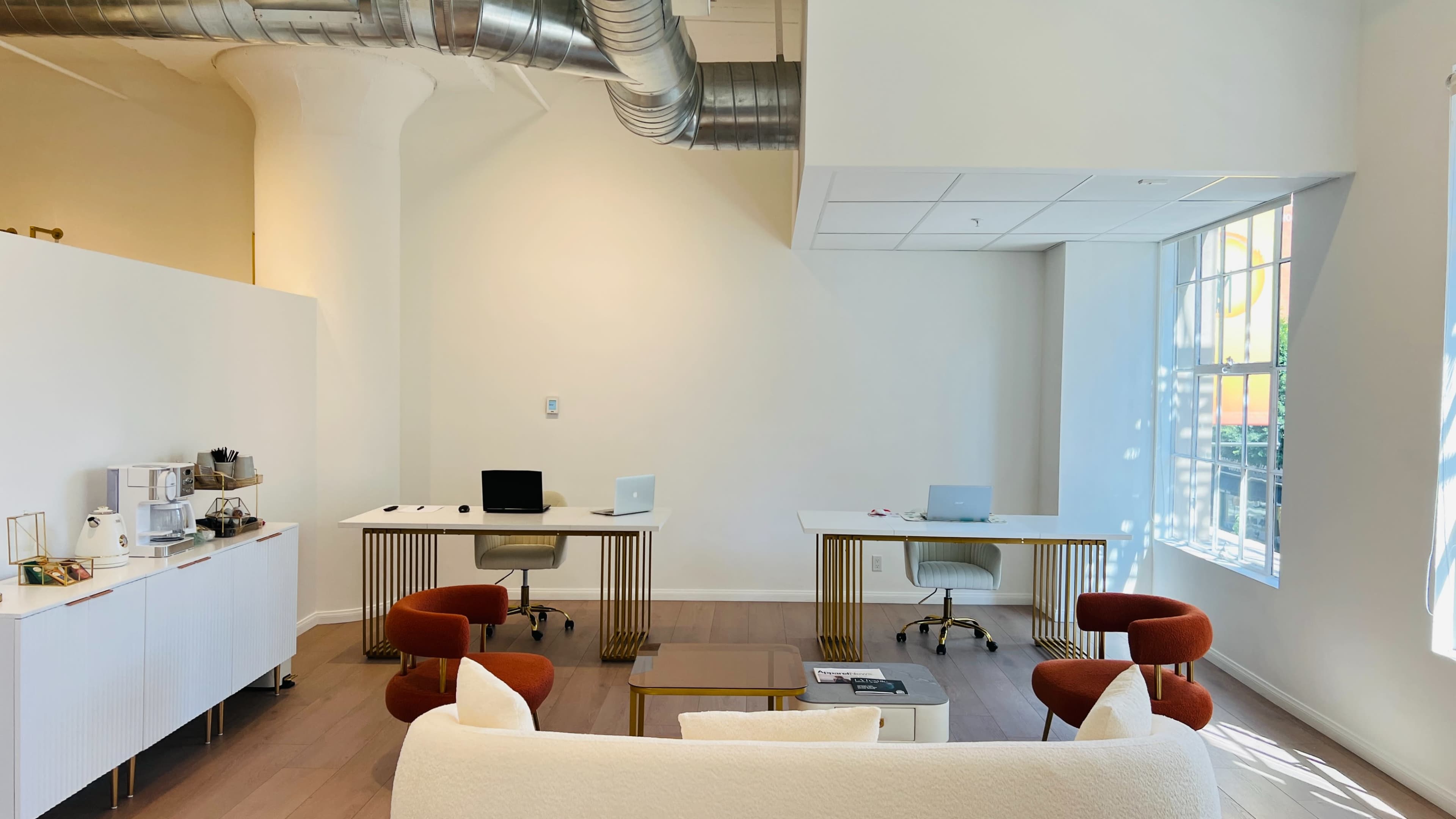 The image shows a modern office space with two desks equipped with computers and a seating area featuring a sofa and two chairs.