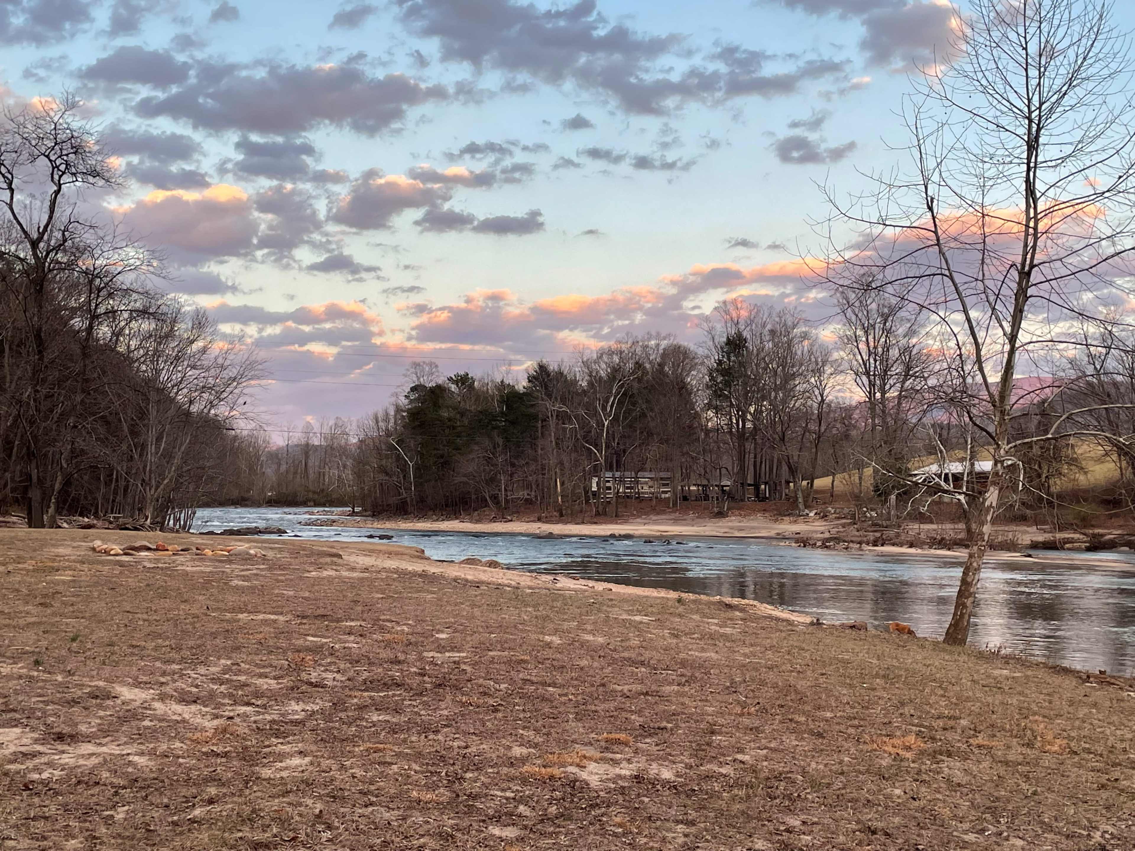 A tranquil river flows alongside a sandy bank, with trees and a house visible in the background under a cloudy sky at sunset.