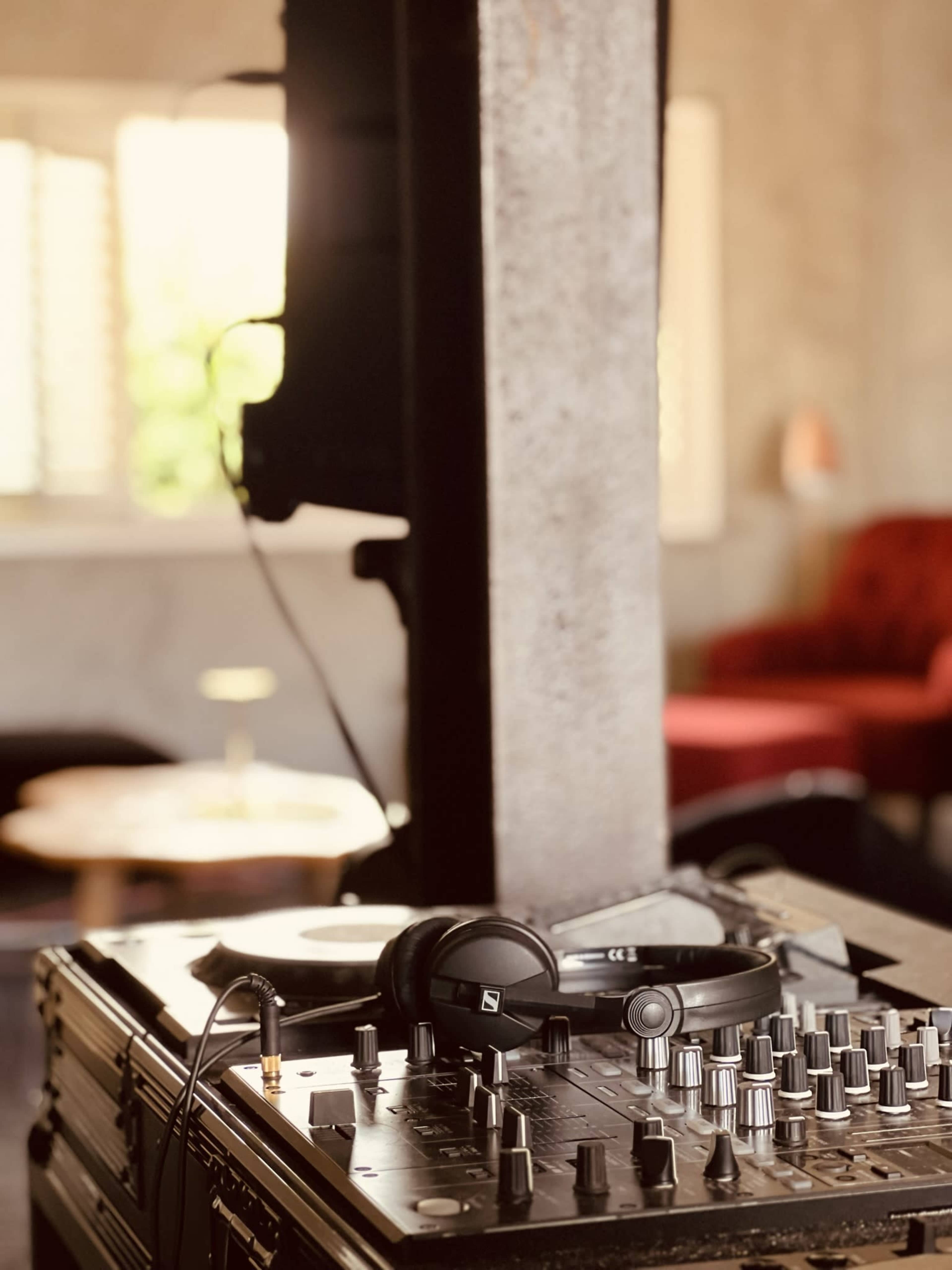 A DJ mixer and headphones are set on a table in a room with a large window and a red chair in the background.