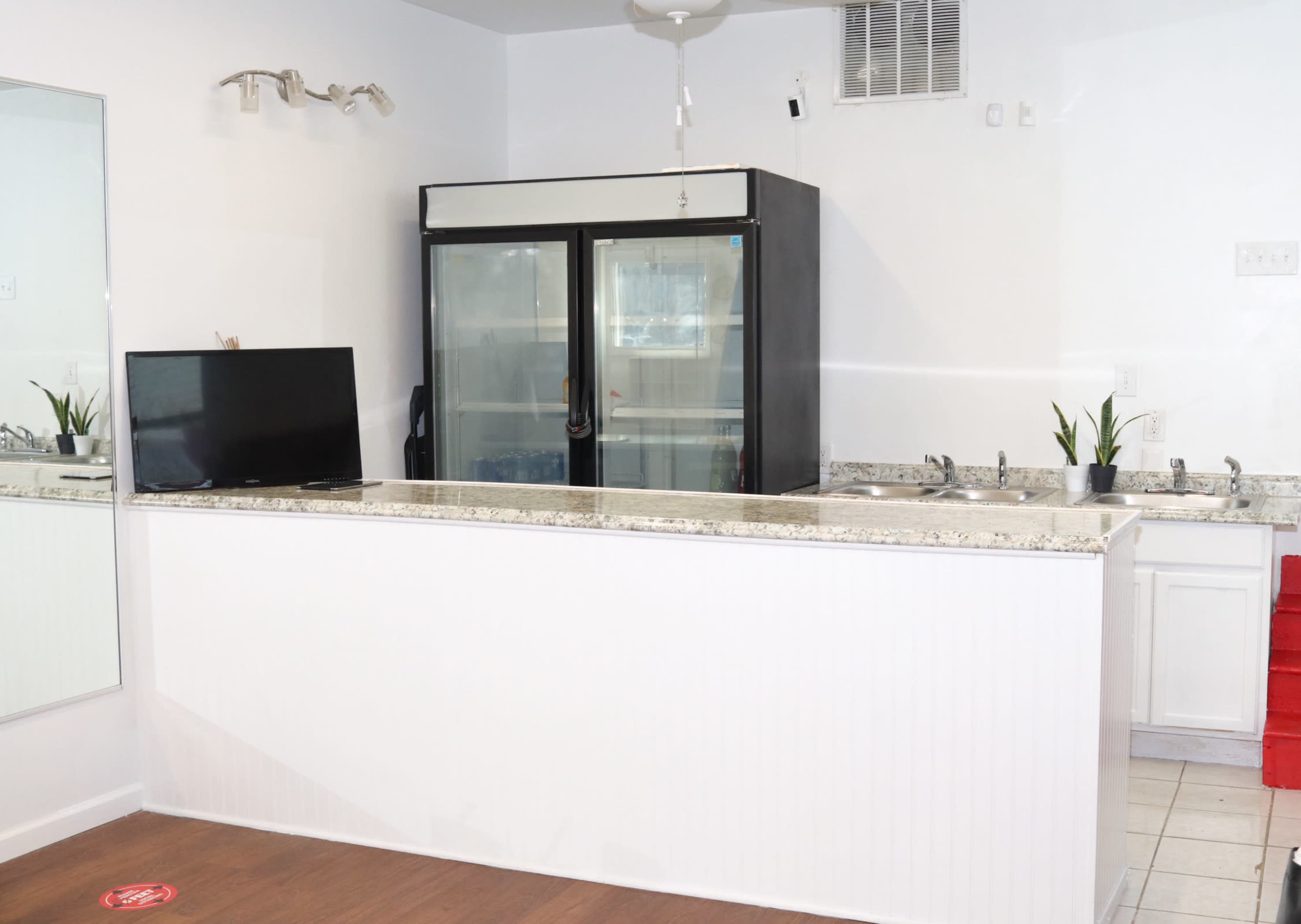 A service counter with a granite top, a wall-mounted television, a black refrigerator, and a small sink, set in a brightly lit space with white walls and a tiled floor.