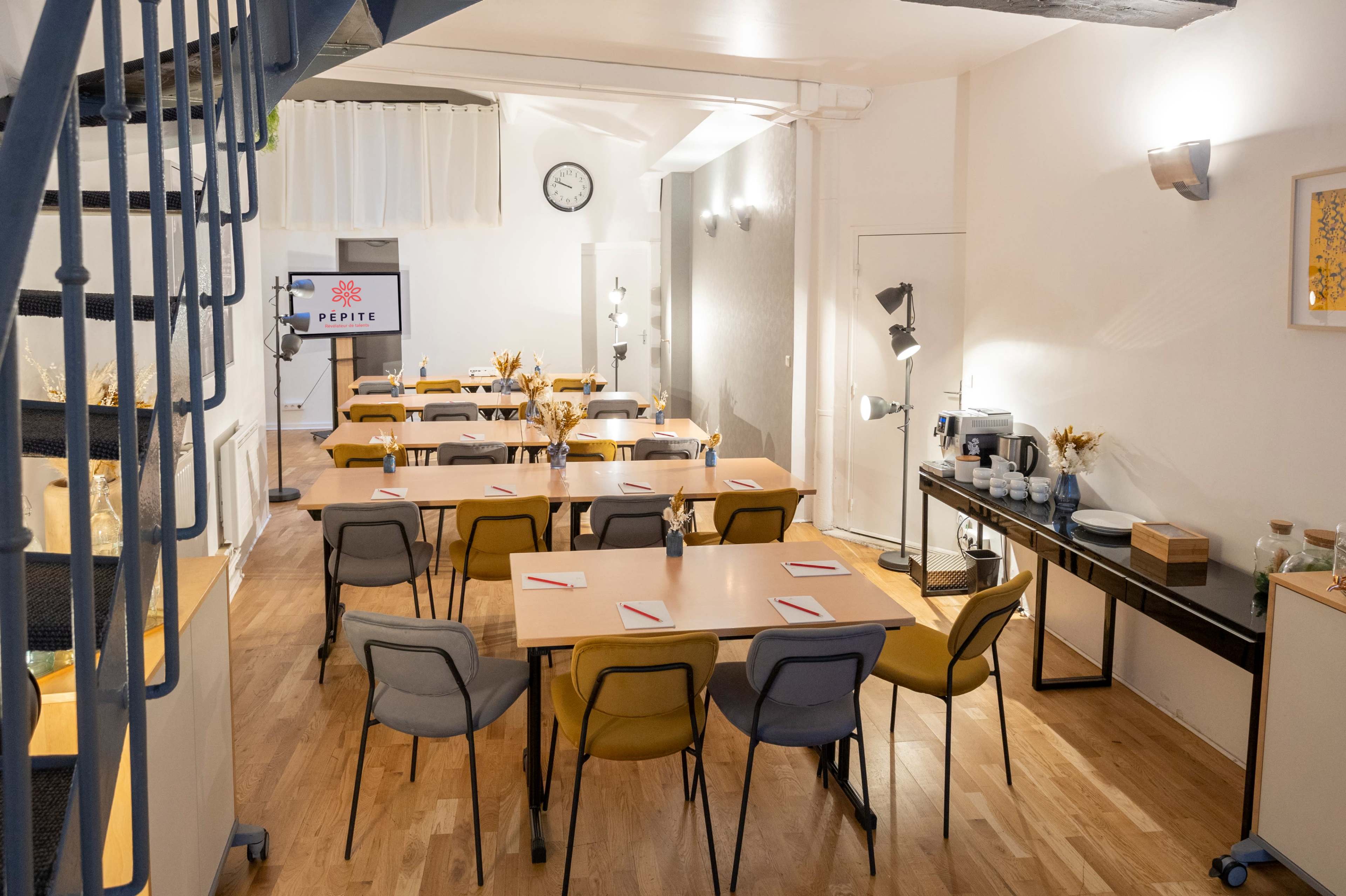 A spacious meeting room features several tables arranged in rows, with chairs and a presentation screen at the front.