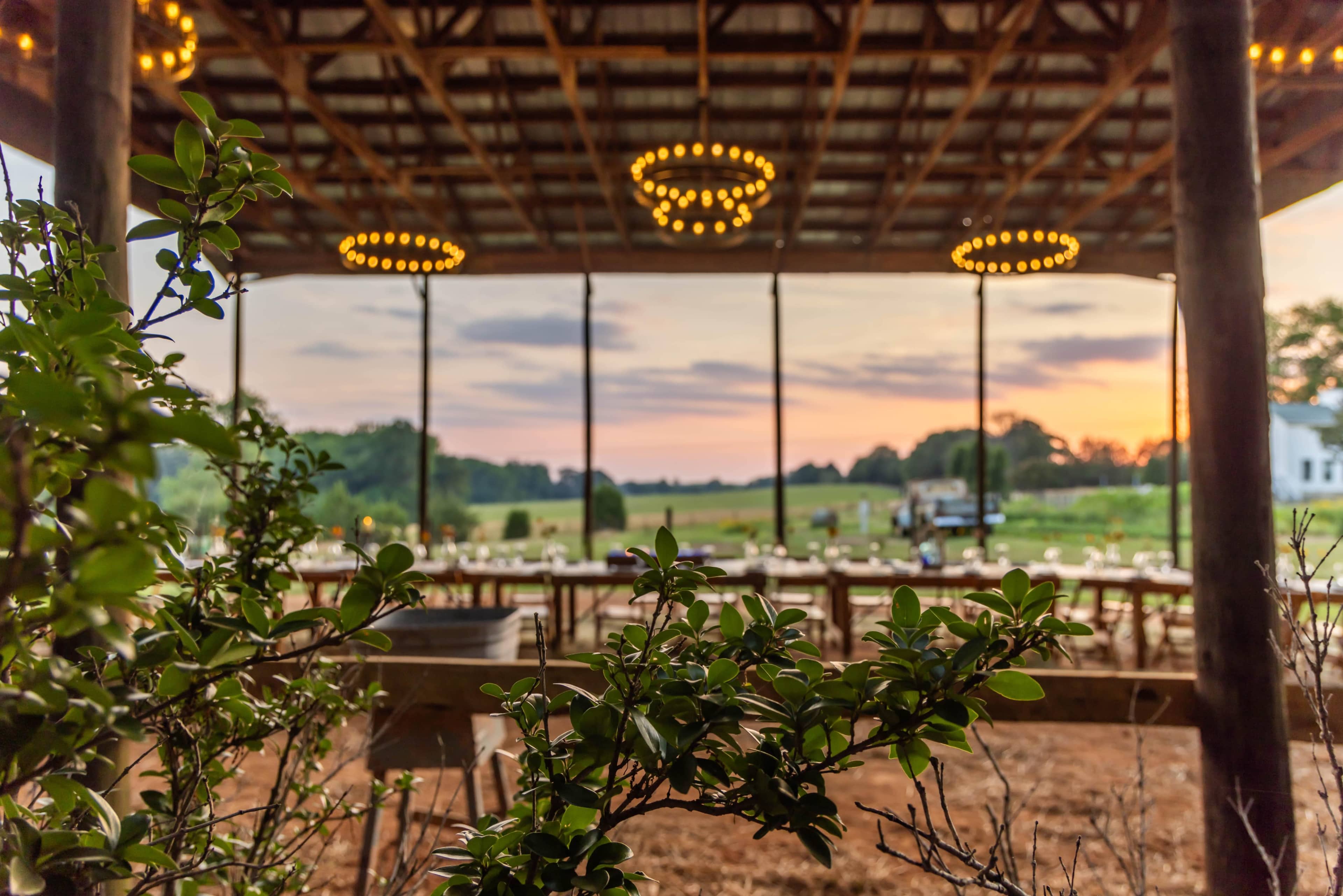 The image shows a spacious outdoor venue with rows of rustic tables set for an event, framed by greenery and illuminated by hanging lights, against a backdrop of a sunset.