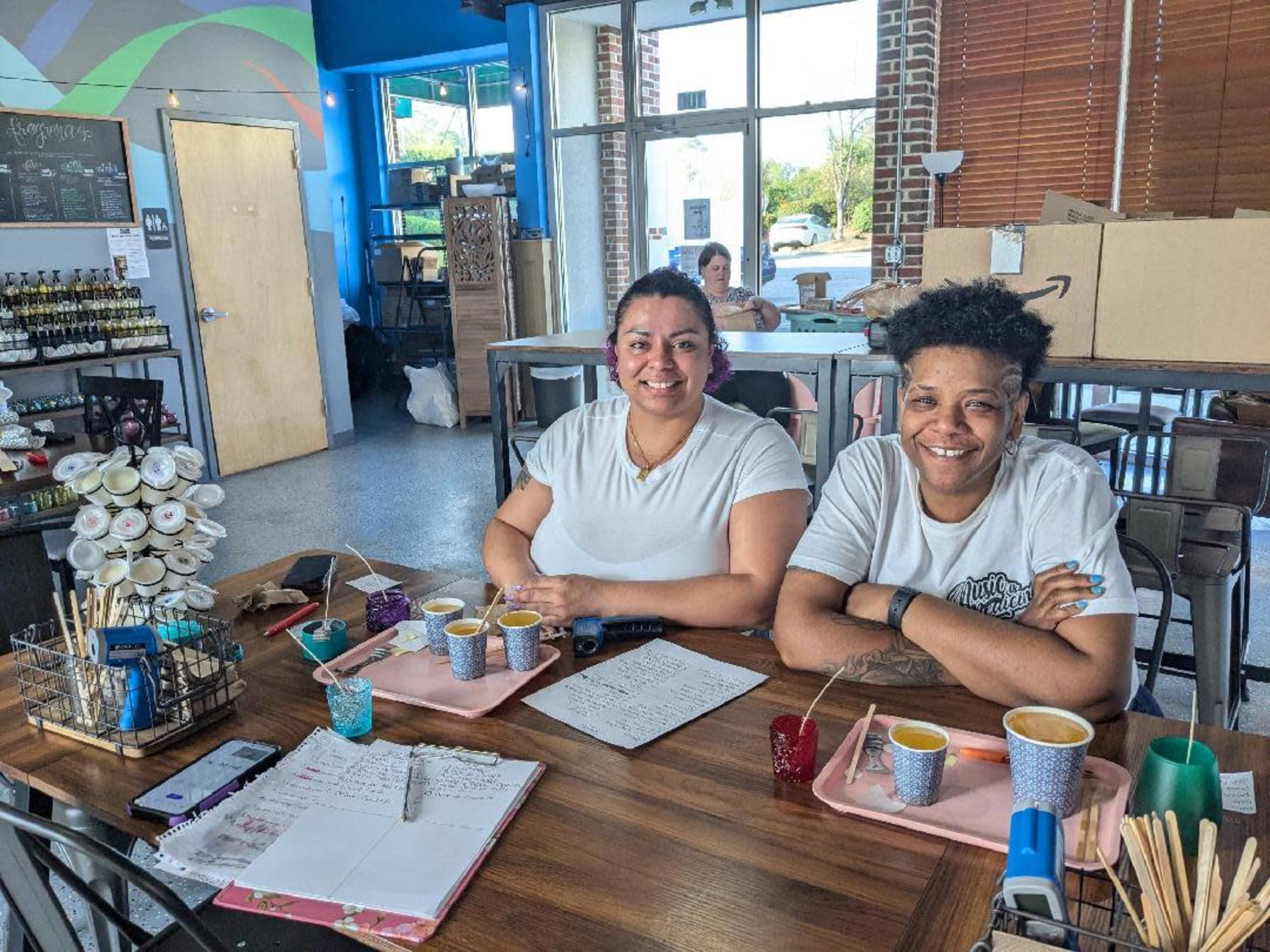 Two women sit at a wooden table in a café, smiling while surrounded by cups and notes.