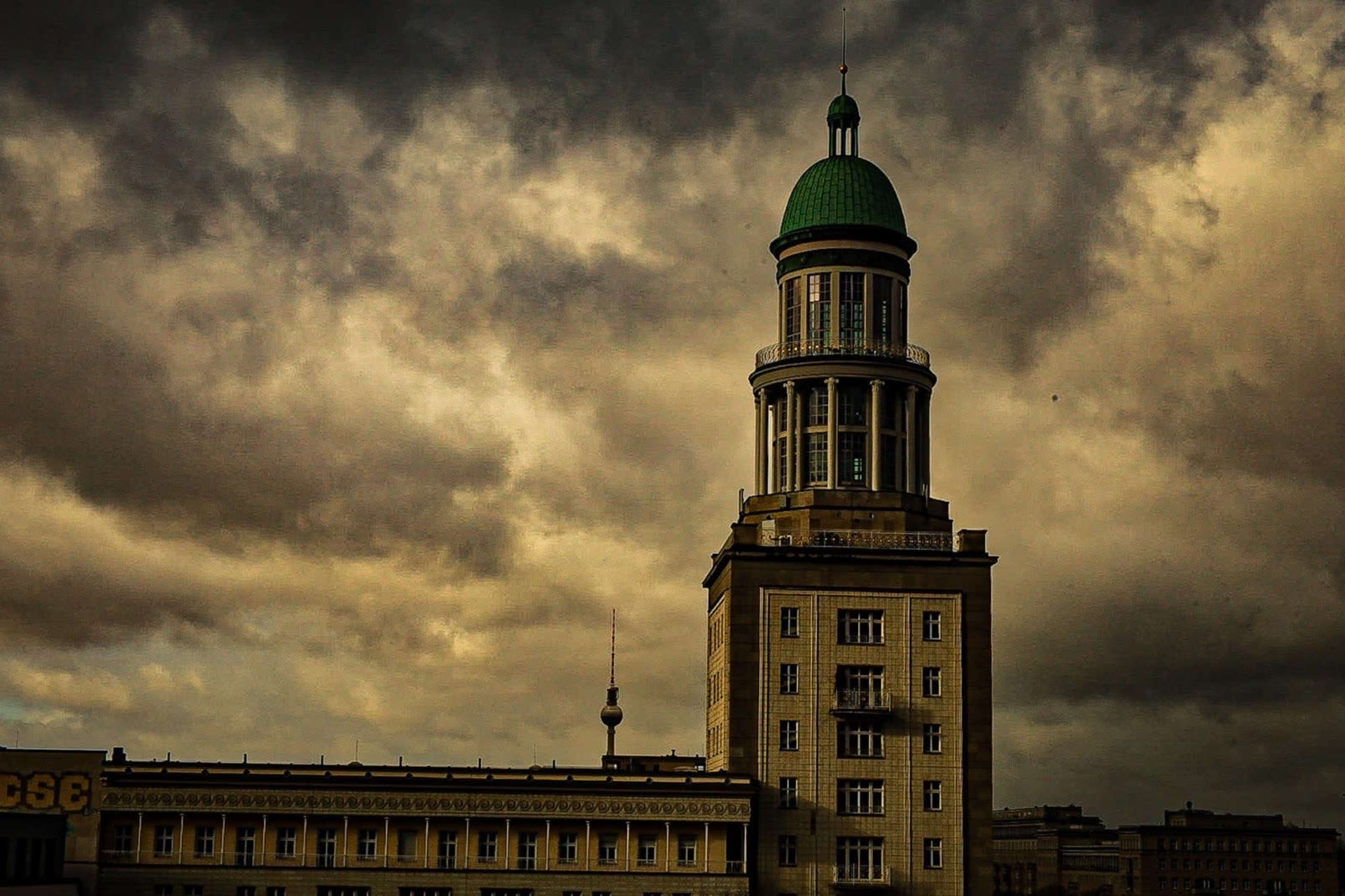 A tall building with a green dome and a clock tower stands under a cloudy sky.