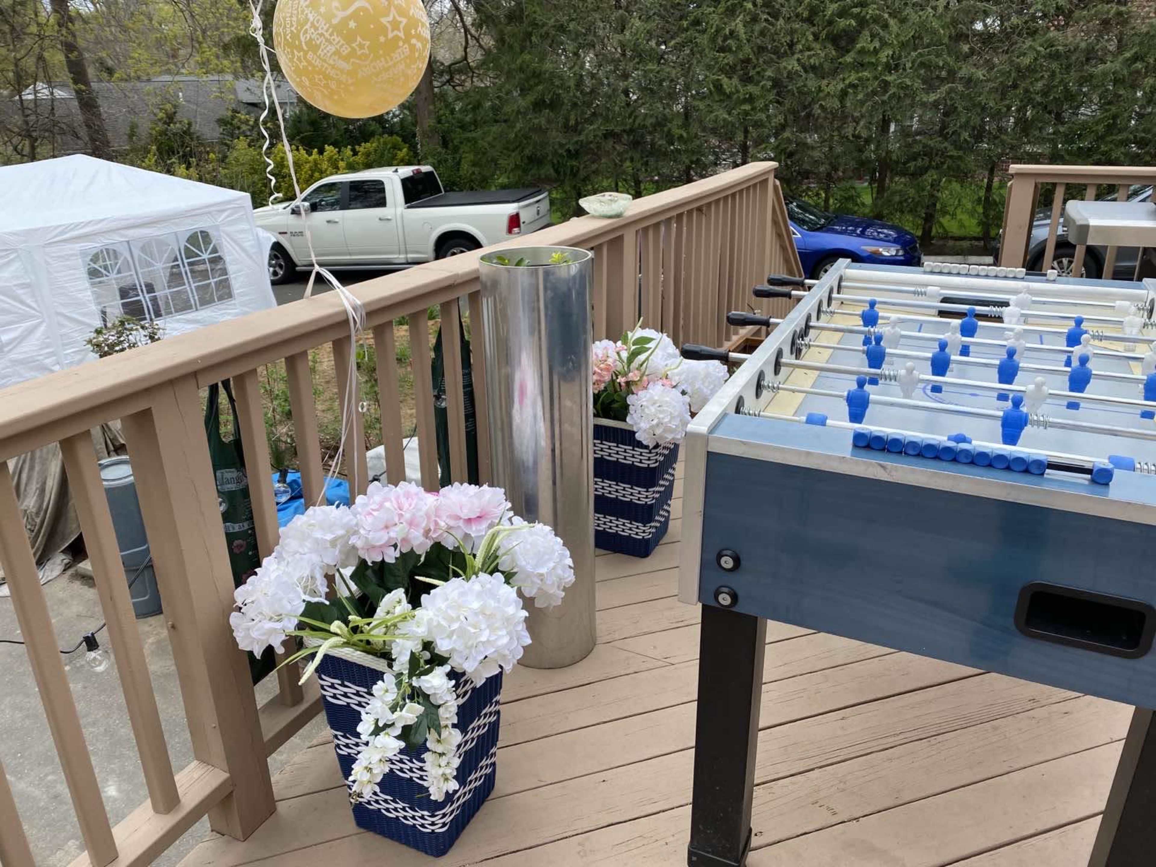 A deck with a foosball table and two decorative planters filled with flowers, alongside a parked truck in the background.
