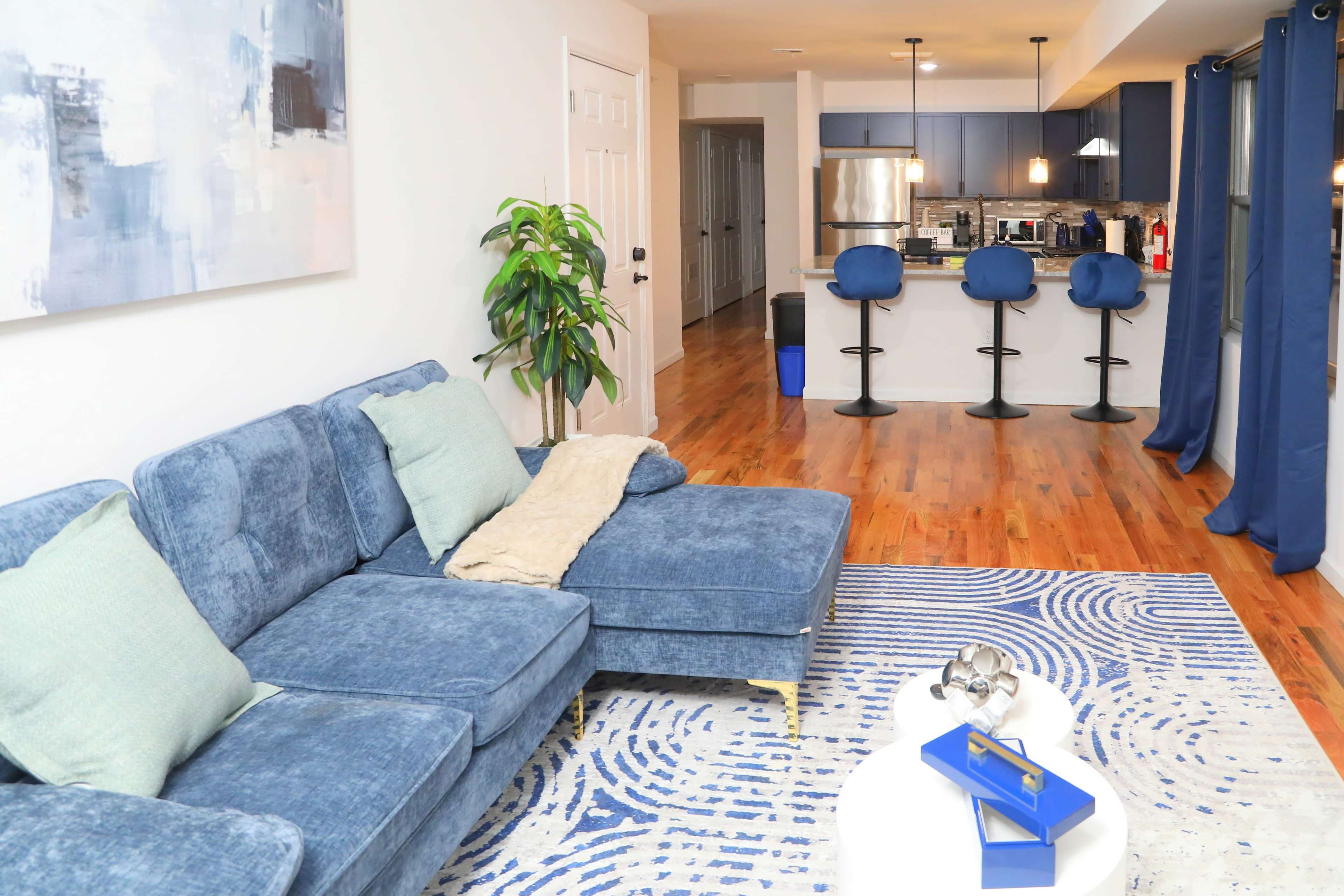 The image shows a modern living room with a blue sectional sofa, a circular coffee table, and a kitchen area visible in the background.