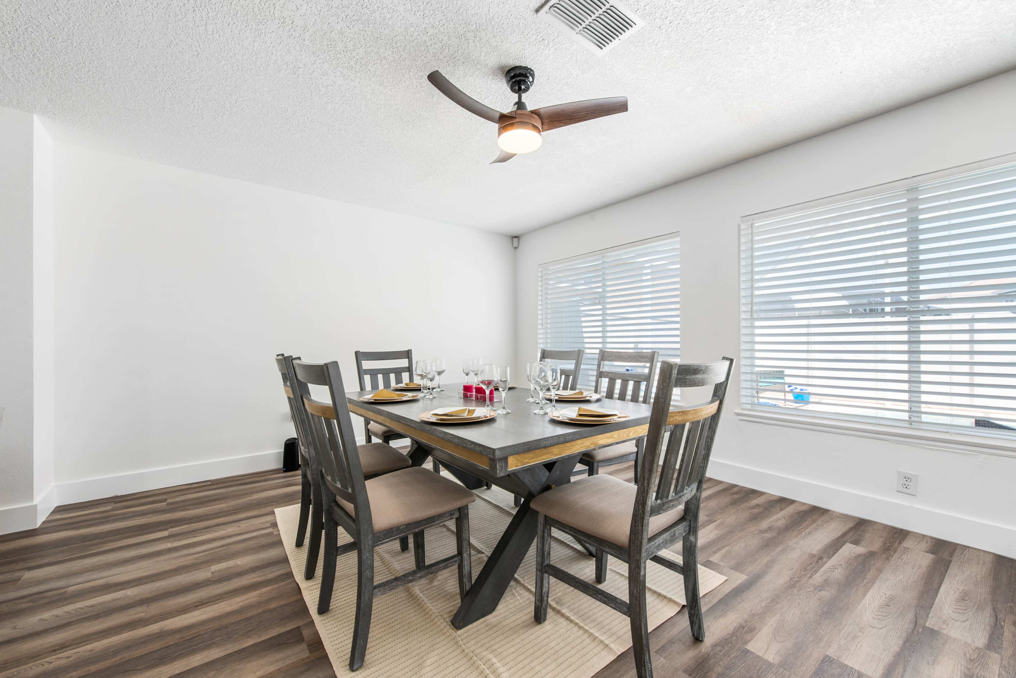 A dining room features a wooden table set for six, surrounded by gray chairs, with ceiling fans and large windows allowing ample natural light.
