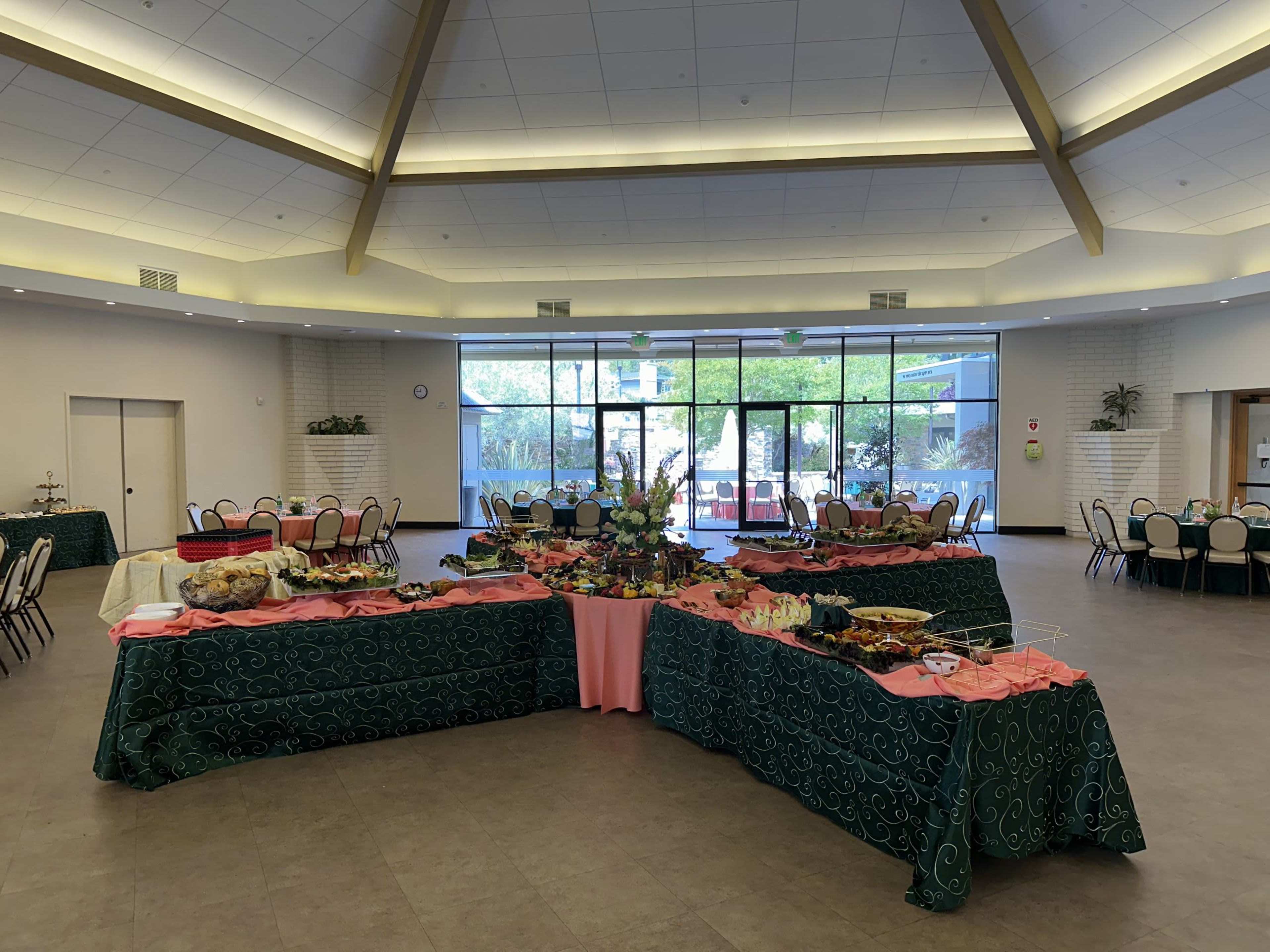 A banquet hall features multiple tables adorned with green tablecloths and pink overlays, arranged in a U-shape, filled with a variety of food items.