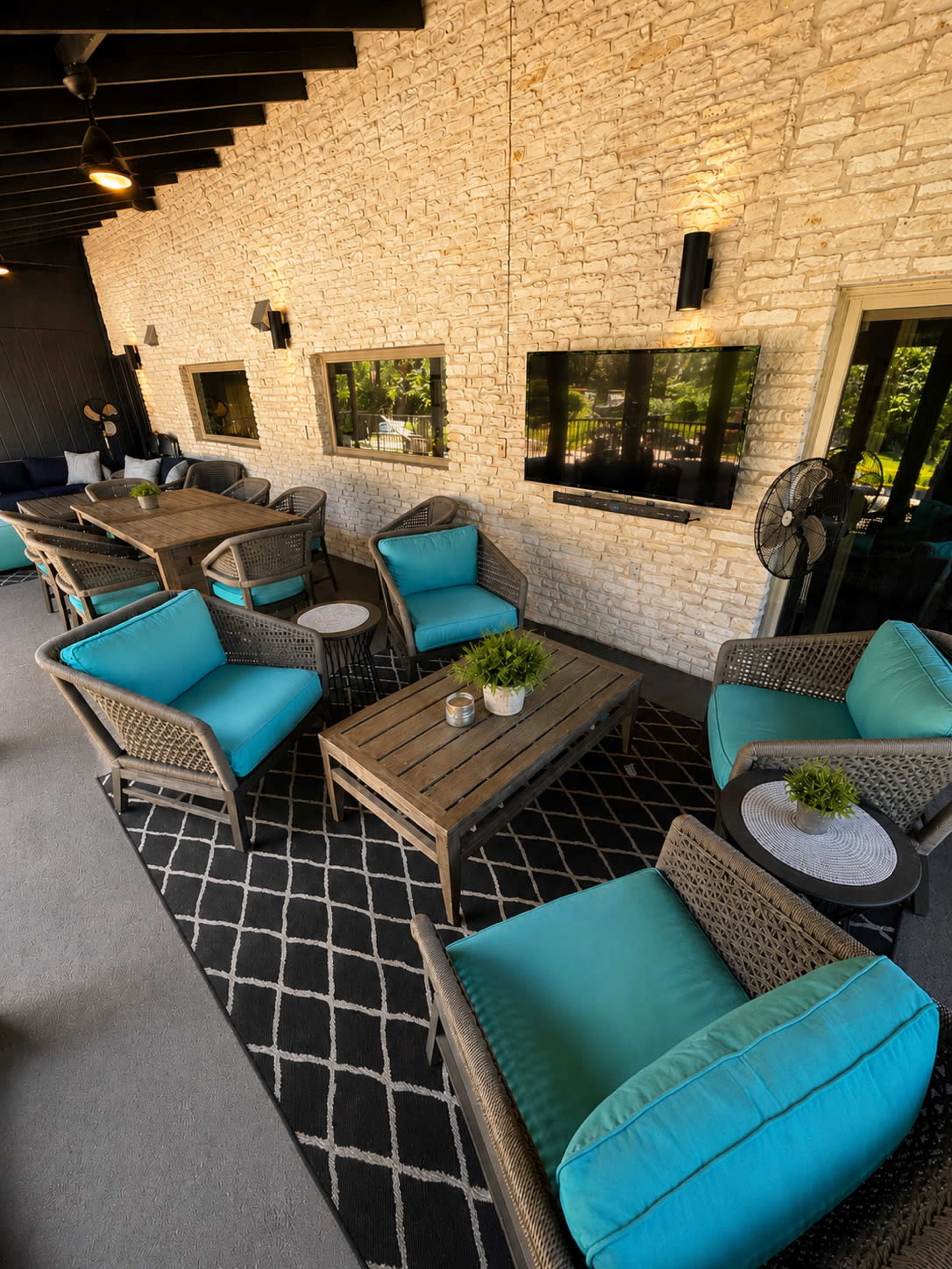 The image shows a cozy outdoor seating area featuring wicker furniture with turquoise cushions, a large wooden table, and stone walls adorned with mounted televisions.