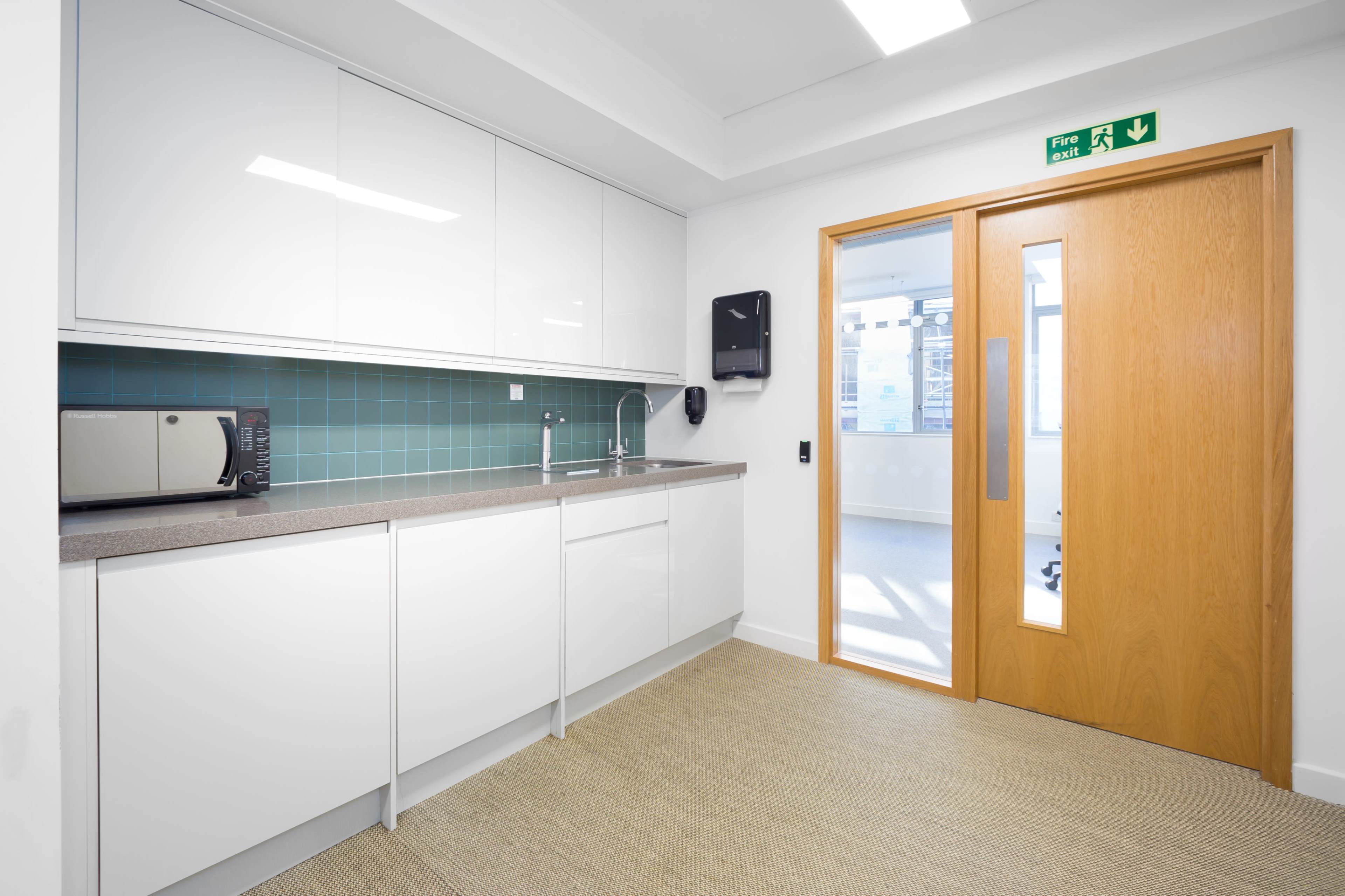 The image shows a modern kitchen area with white cabinets, a sink, a microwave, and a wooden door leading to another room.