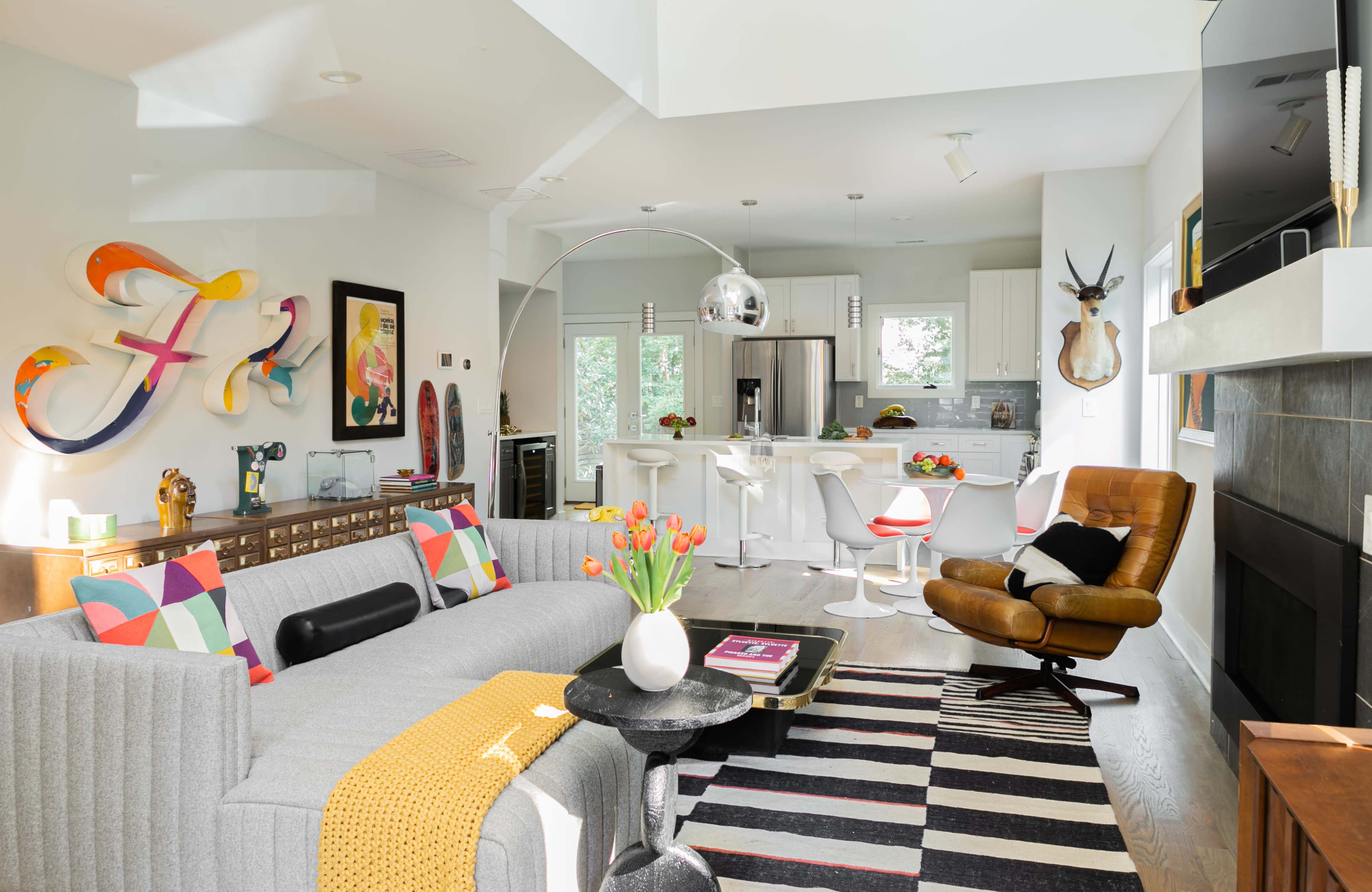 A modern living space features a gray sofa, a black coffee table, colorful decorative pillows, and an open kitchen with white cabinetry and bar stools.