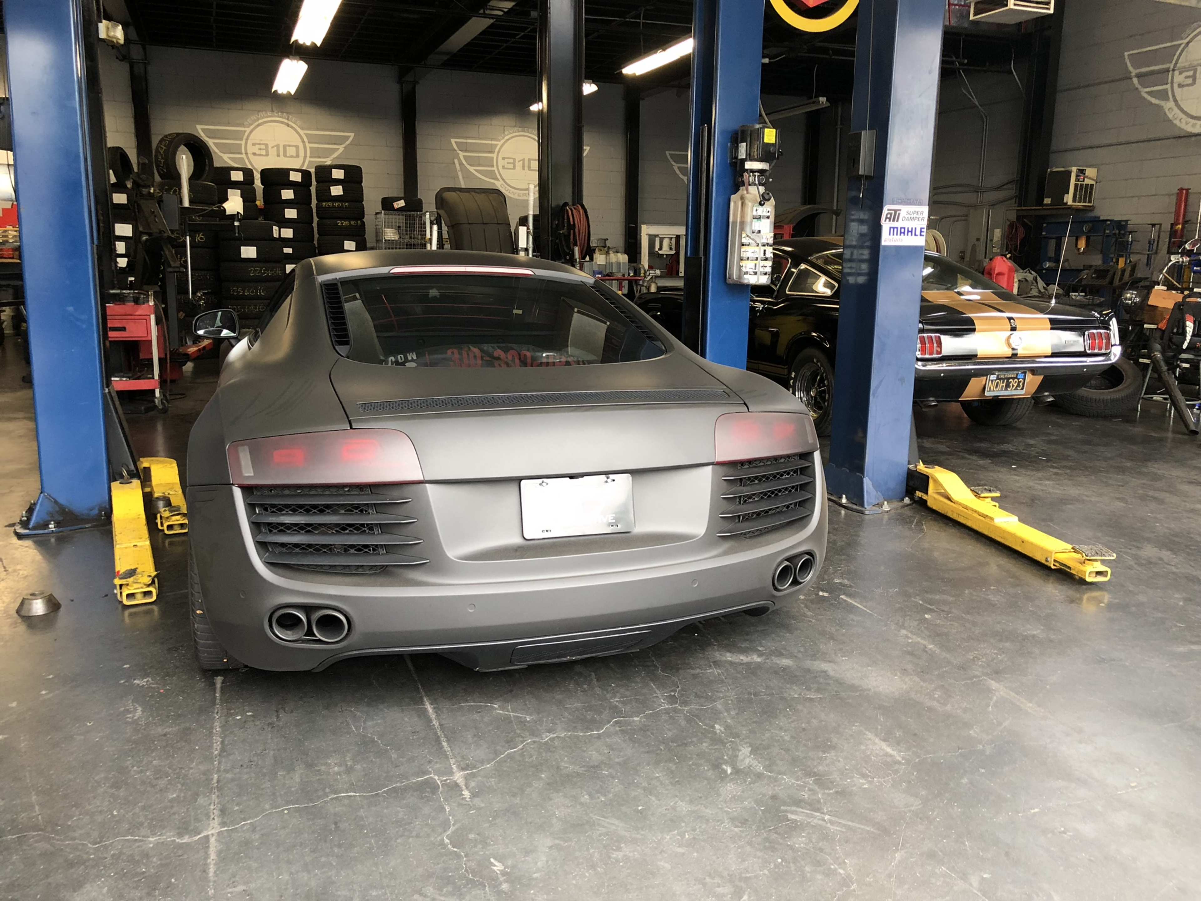 The image shows a matte black Audi R8 and a classic Mustang parked in a garage with car lifts and tools visible in the background.