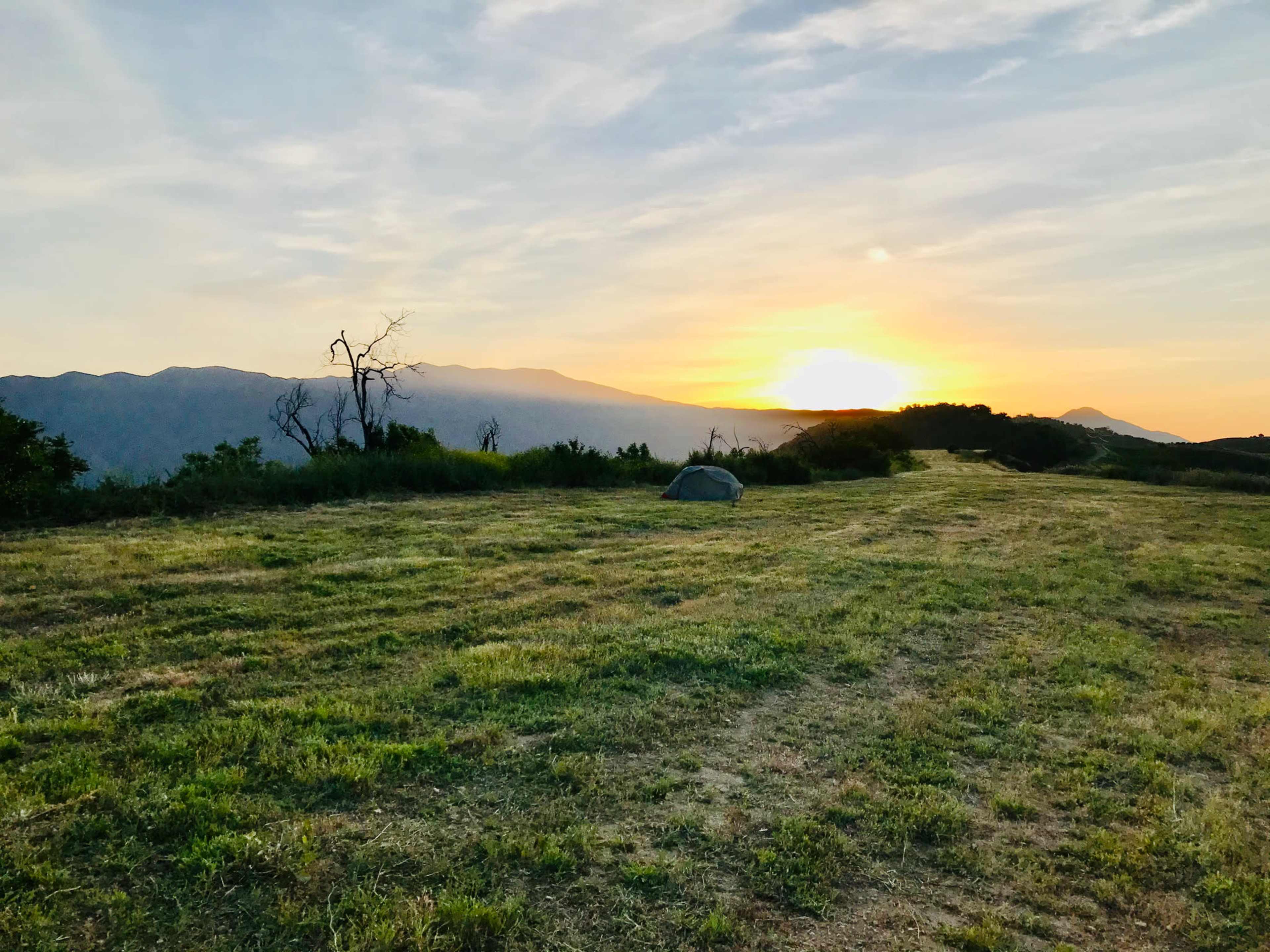 A tent is set up on a grassy field with mountains in the background as the sun rises above the horizon.