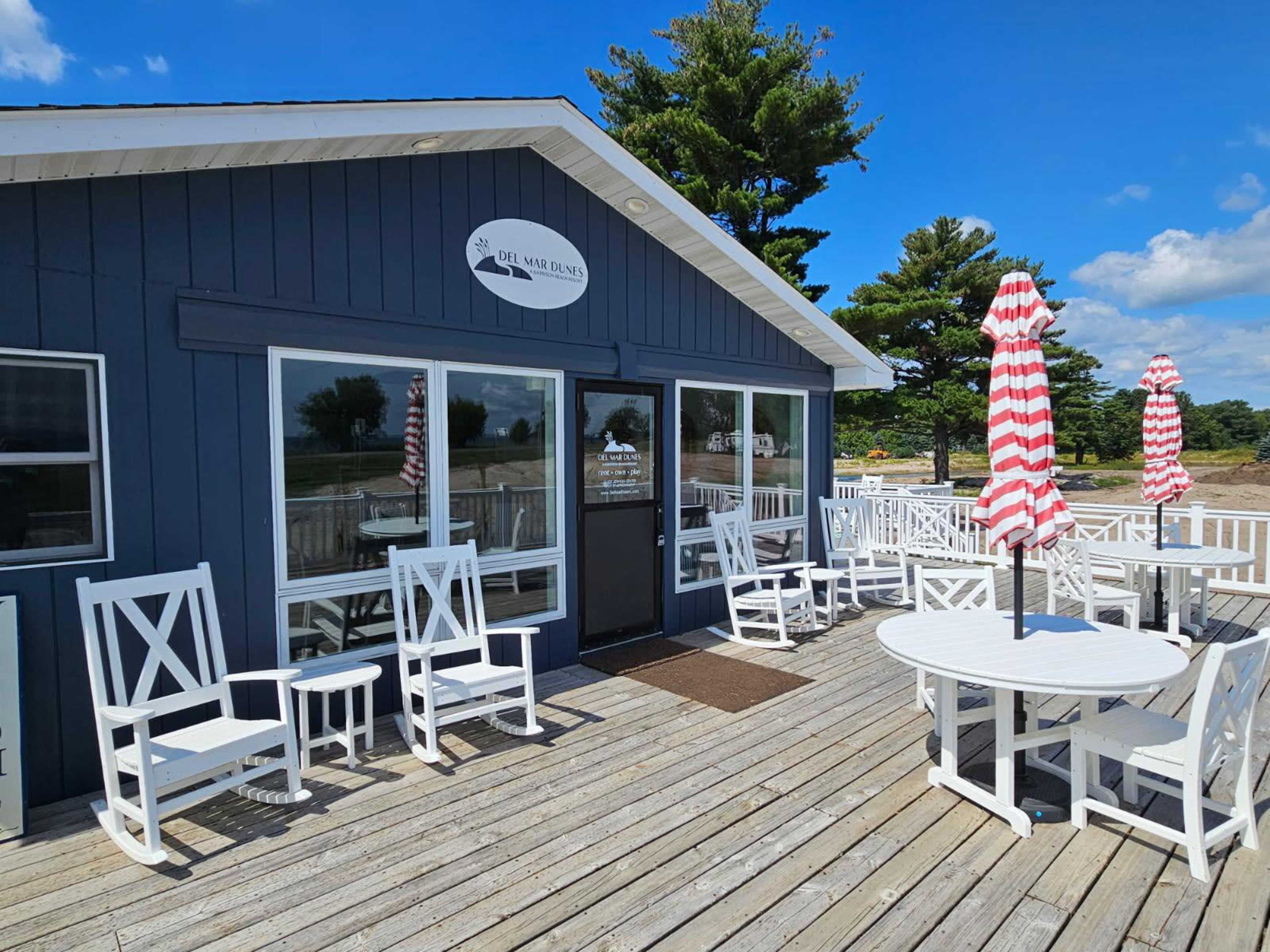 The image shows a blue wooden building with a sign that reads "Del Mar Jones" and outdoor seating featuring white chairs and round tables on a deck surrounded by trees.
