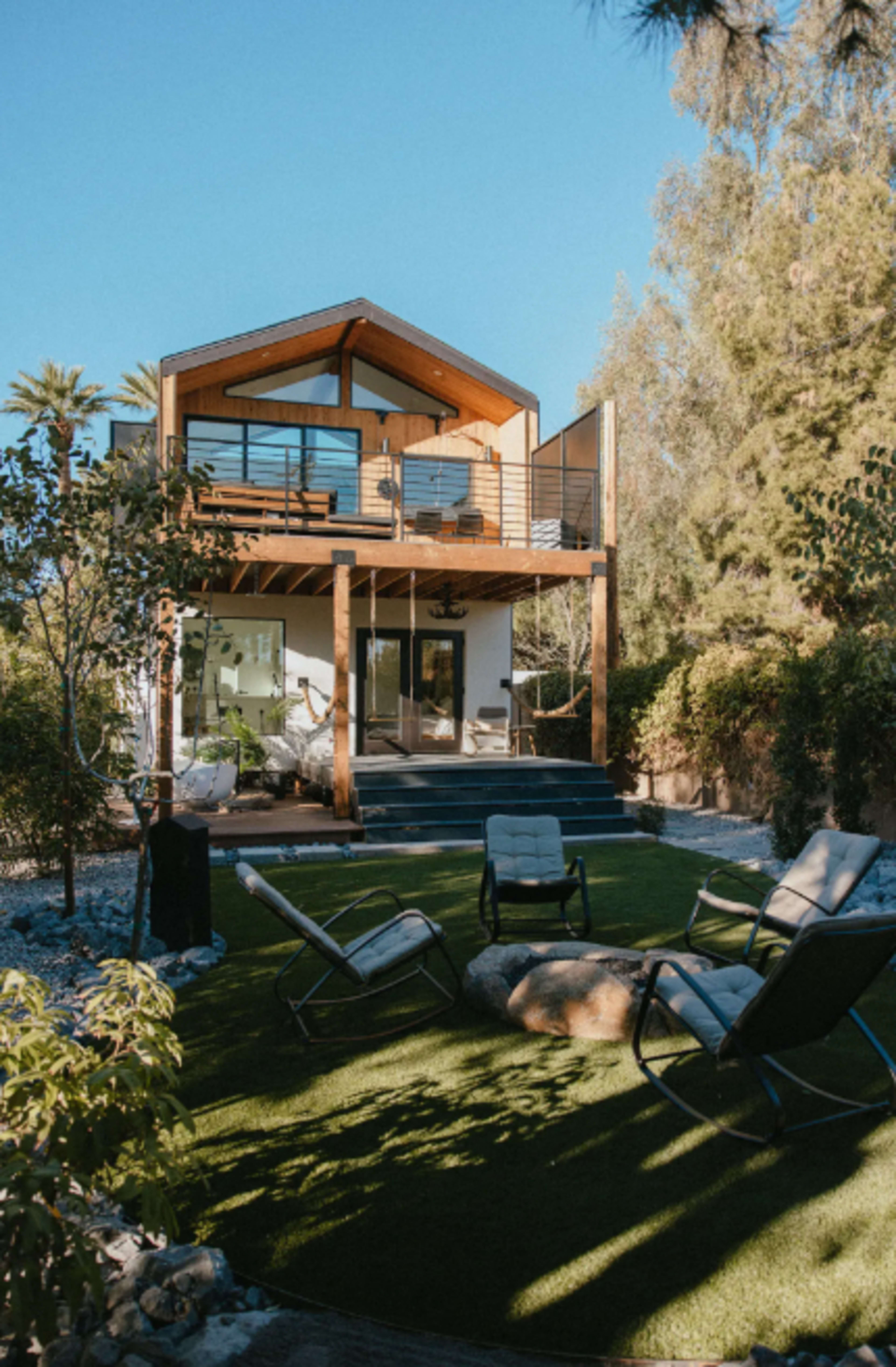 A modern two-story house with a wooden balcony and steps, surrounded by greenery and outdoor seating.