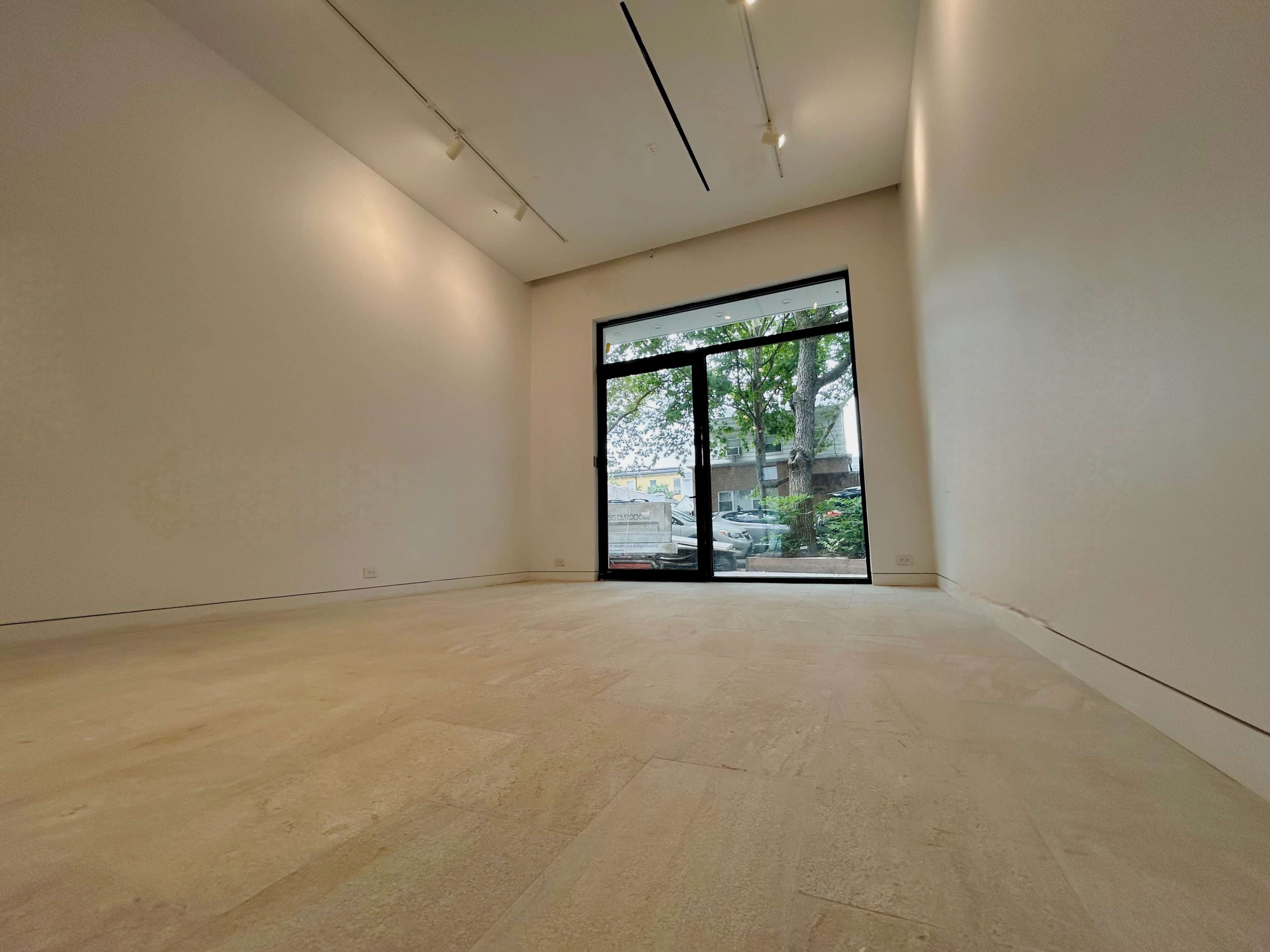 The image shows a minimalist gallery space with a large glass window letting in natural light and an empty floor.