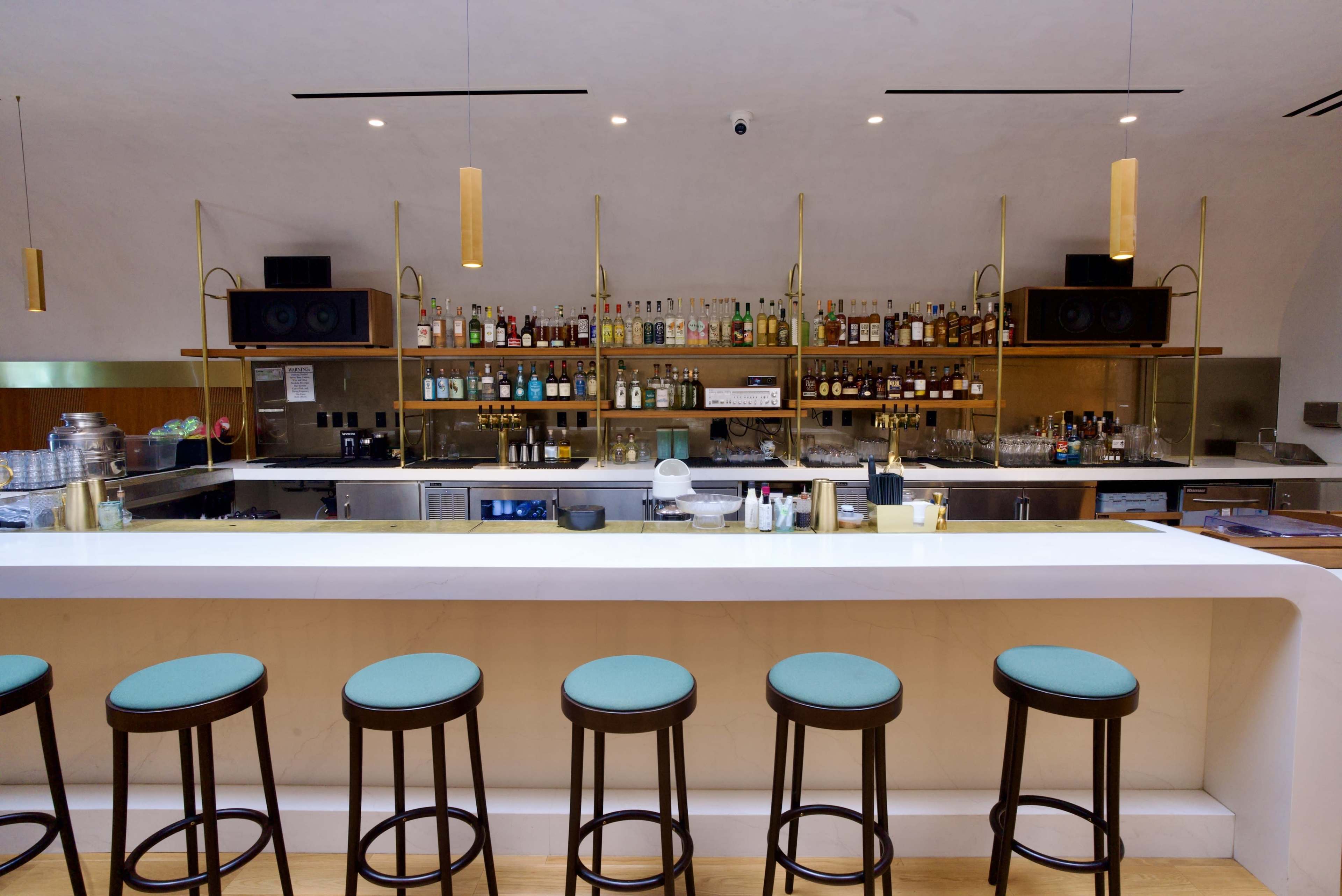 The image shows a modern bar with a long white counter, several high stools, and shelves stocked with various bottles of alcohol.