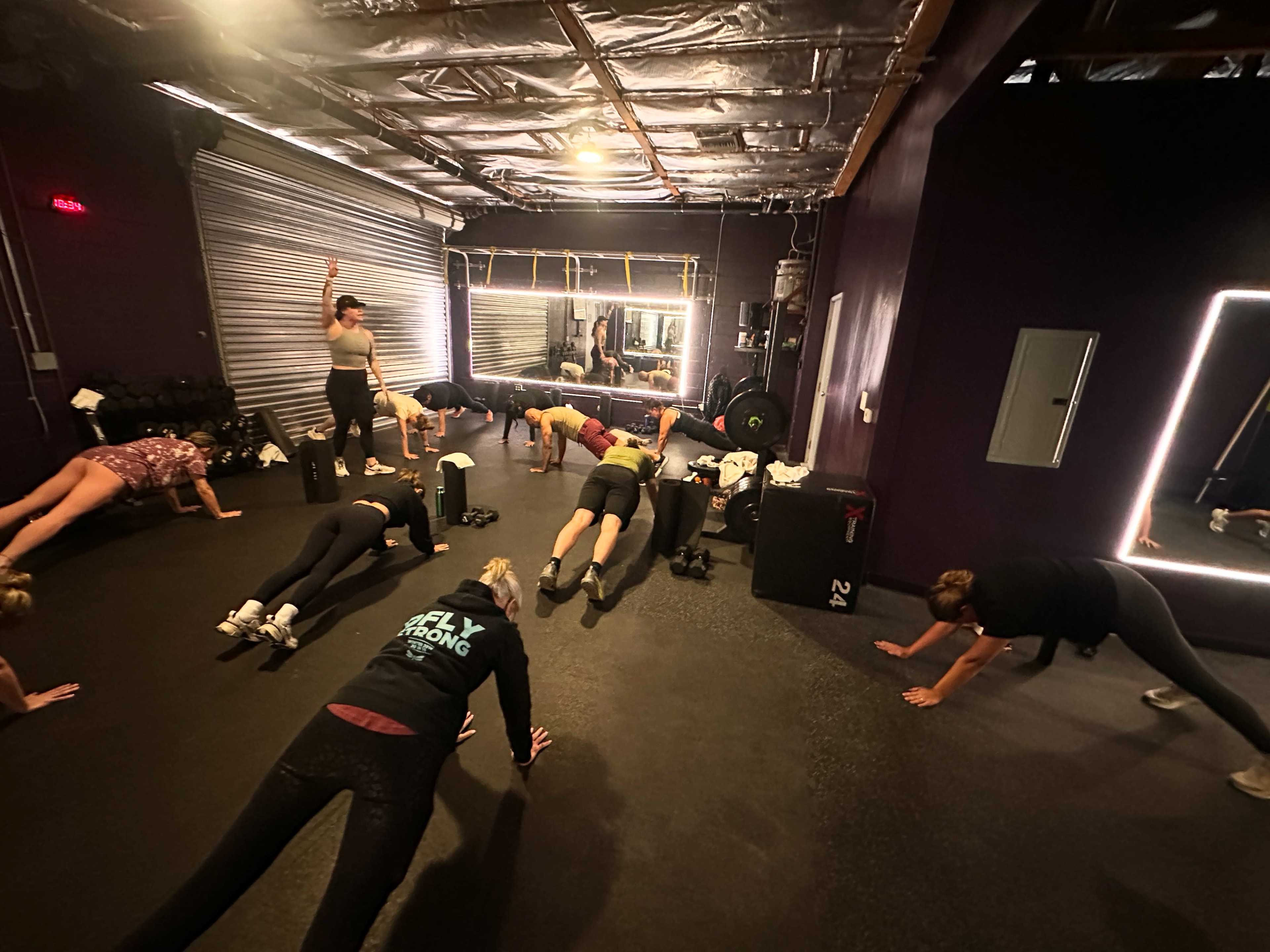 A group of people participates in a fitness class, performing plank exercises on mats in a gym setting.