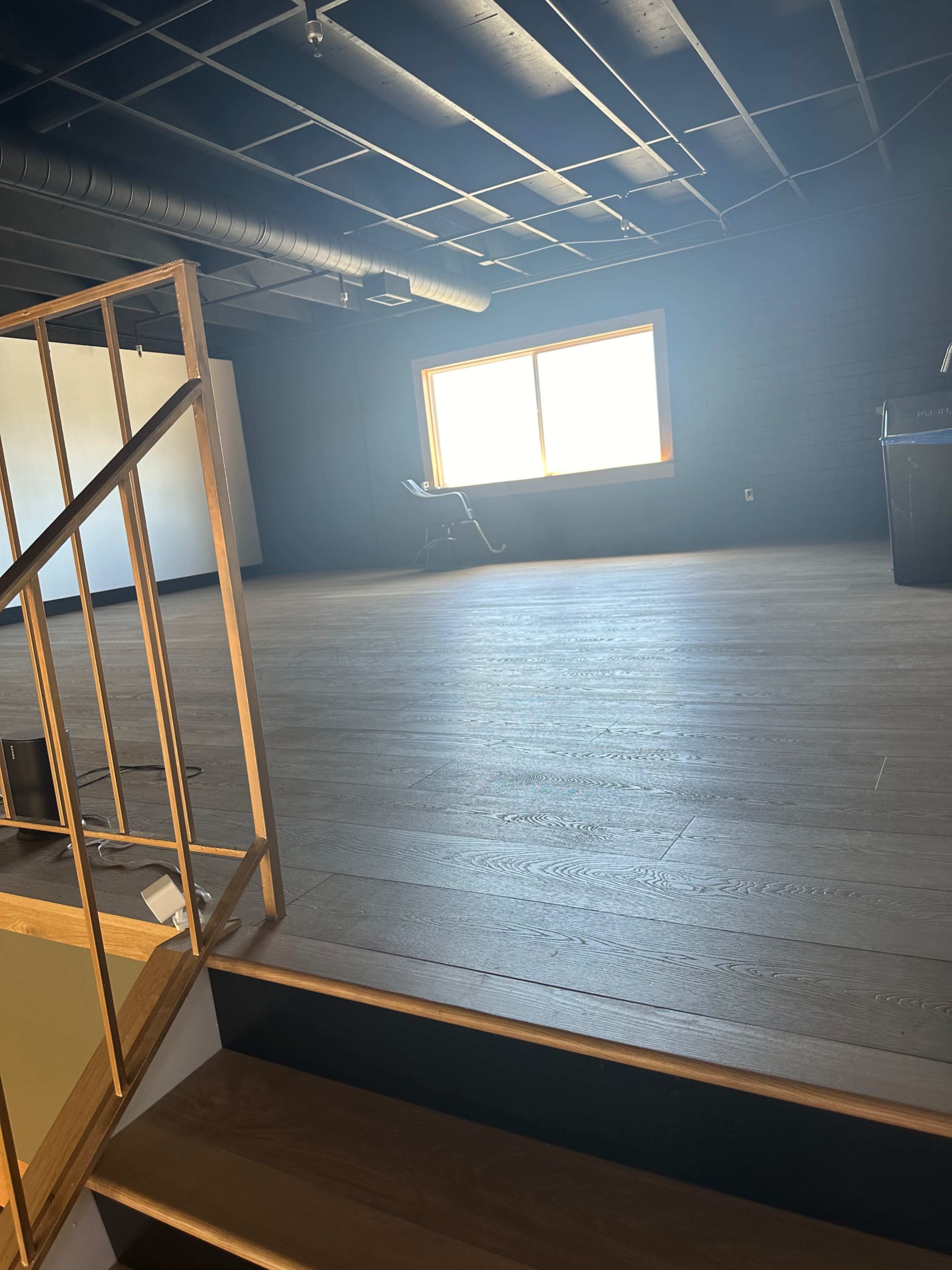 The image shows a spacious, empty room with wooden flooring and a large window allowing natural light, viewed from a staircase.