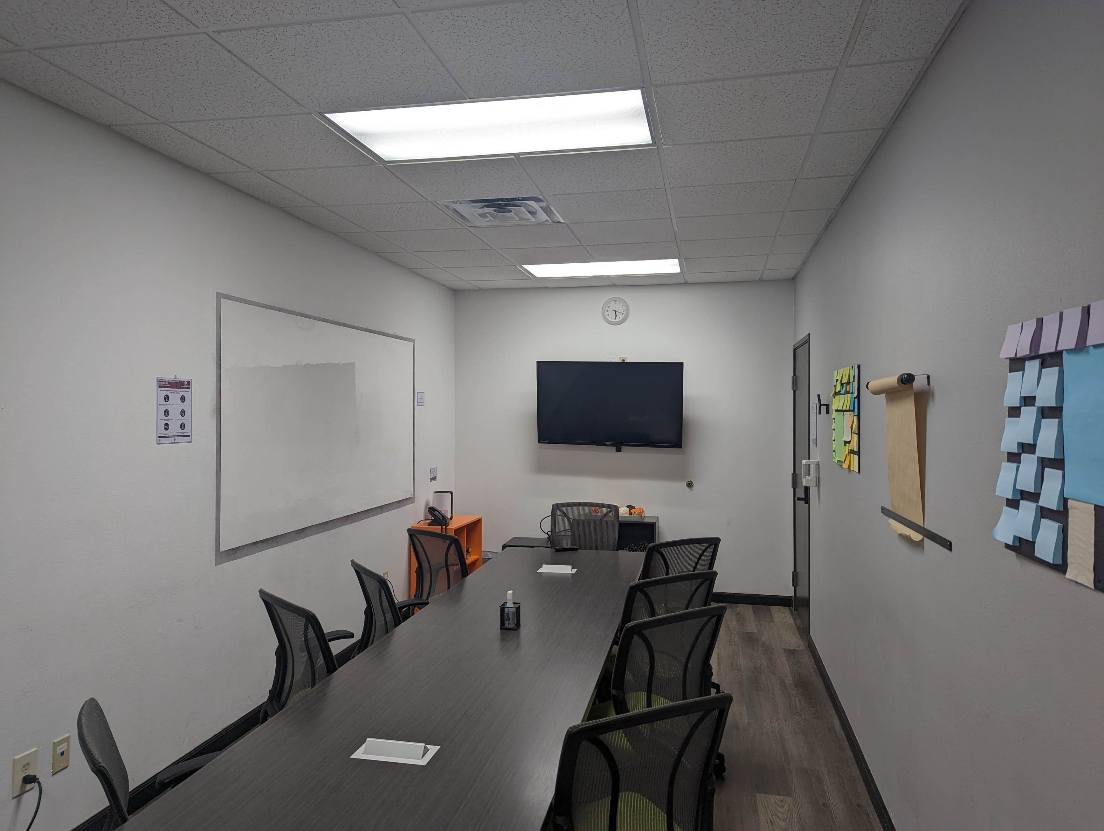 A conference room features a long table surrounded by chairs, a whiteboard, a wall-mounted television, and various notes on the walls.