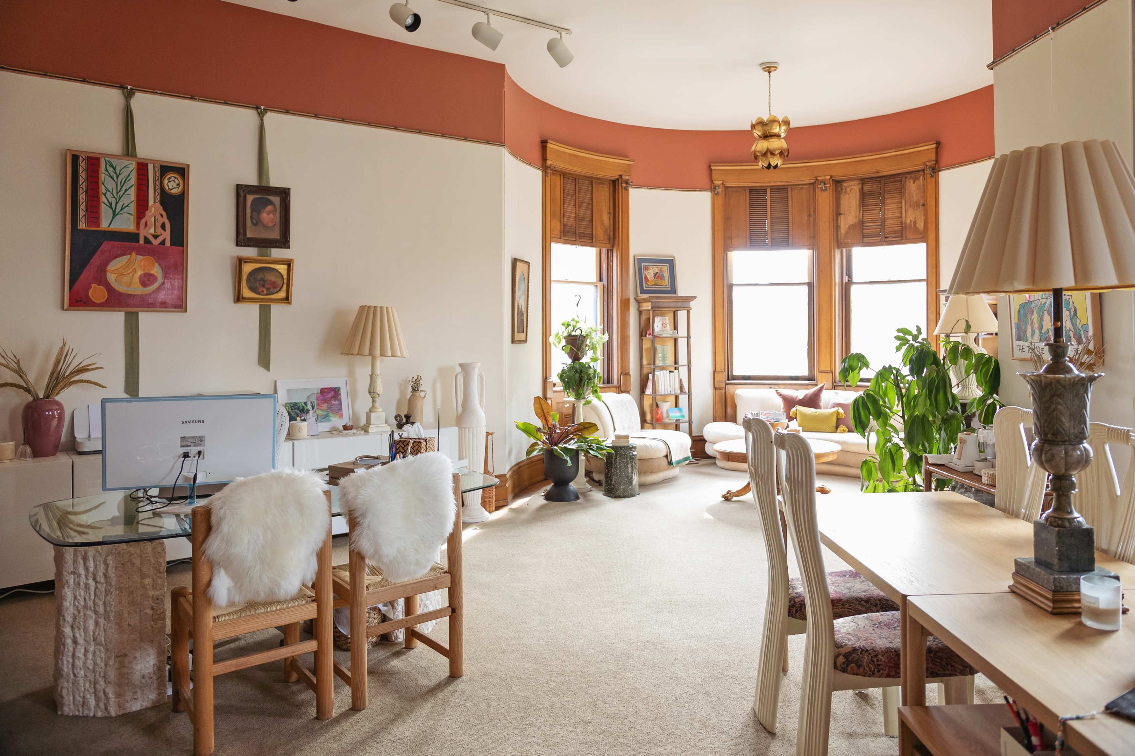 A brightly lit room features a mix of seating areas, a wooden dining table, and decorative plants against a backdrop of large windows.