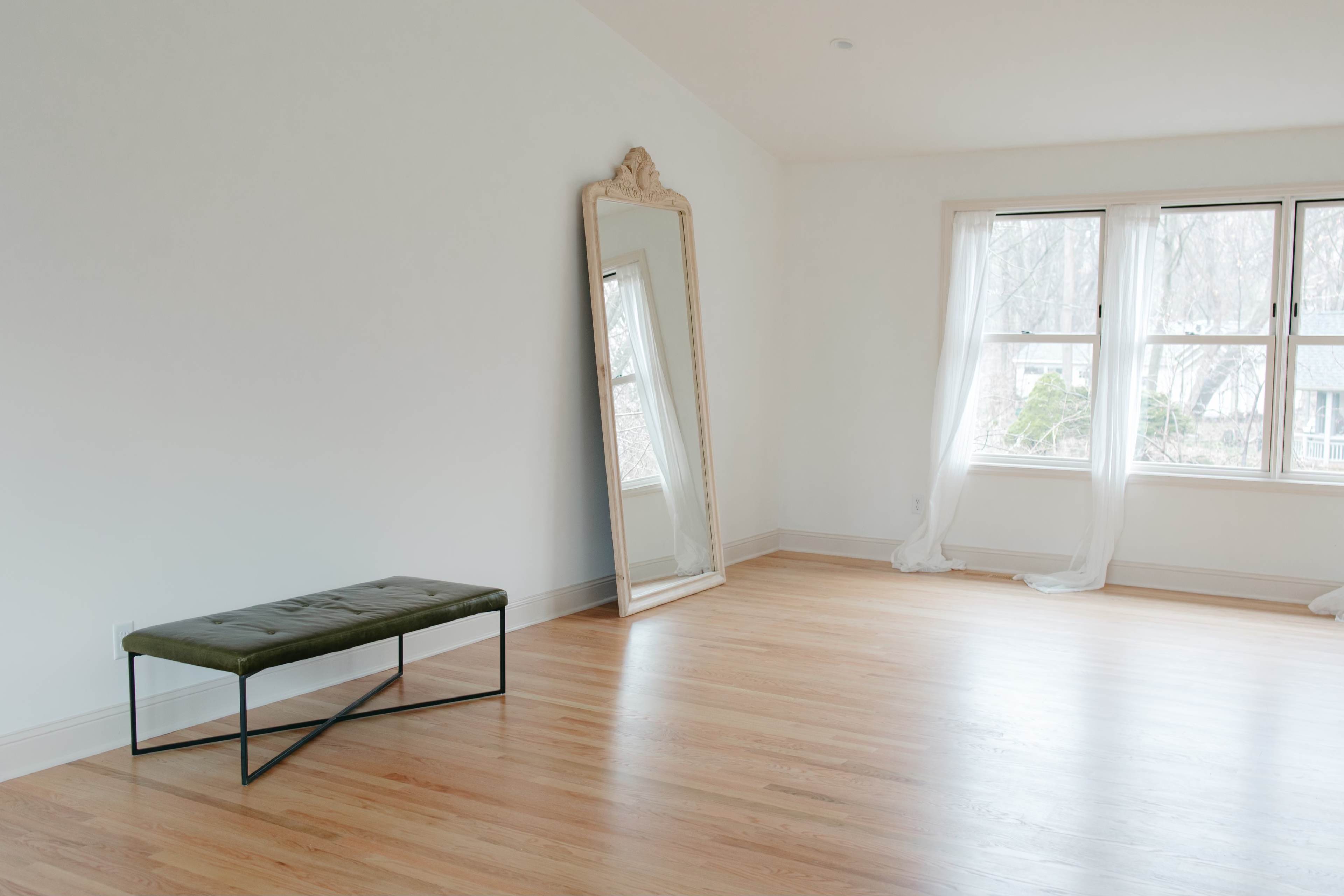 The scene features a sparsely furnished room with a green bench and an ornate mirror against a light-colored wall, illuminated by natural light from large windows.