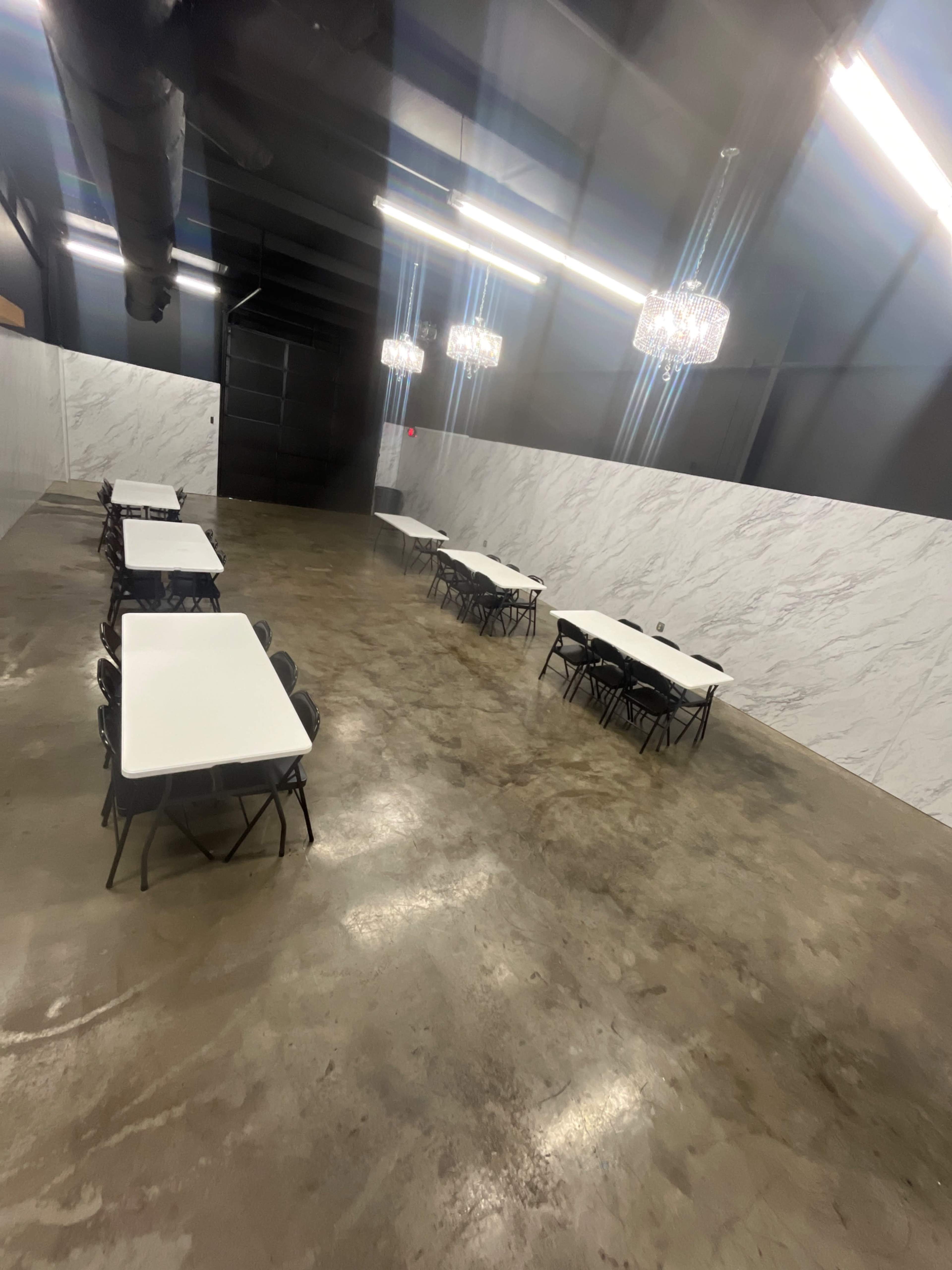 A spacious, empty room with concrete flooring, several white tables set with chairs, and two chandeliers illuminating the space.
