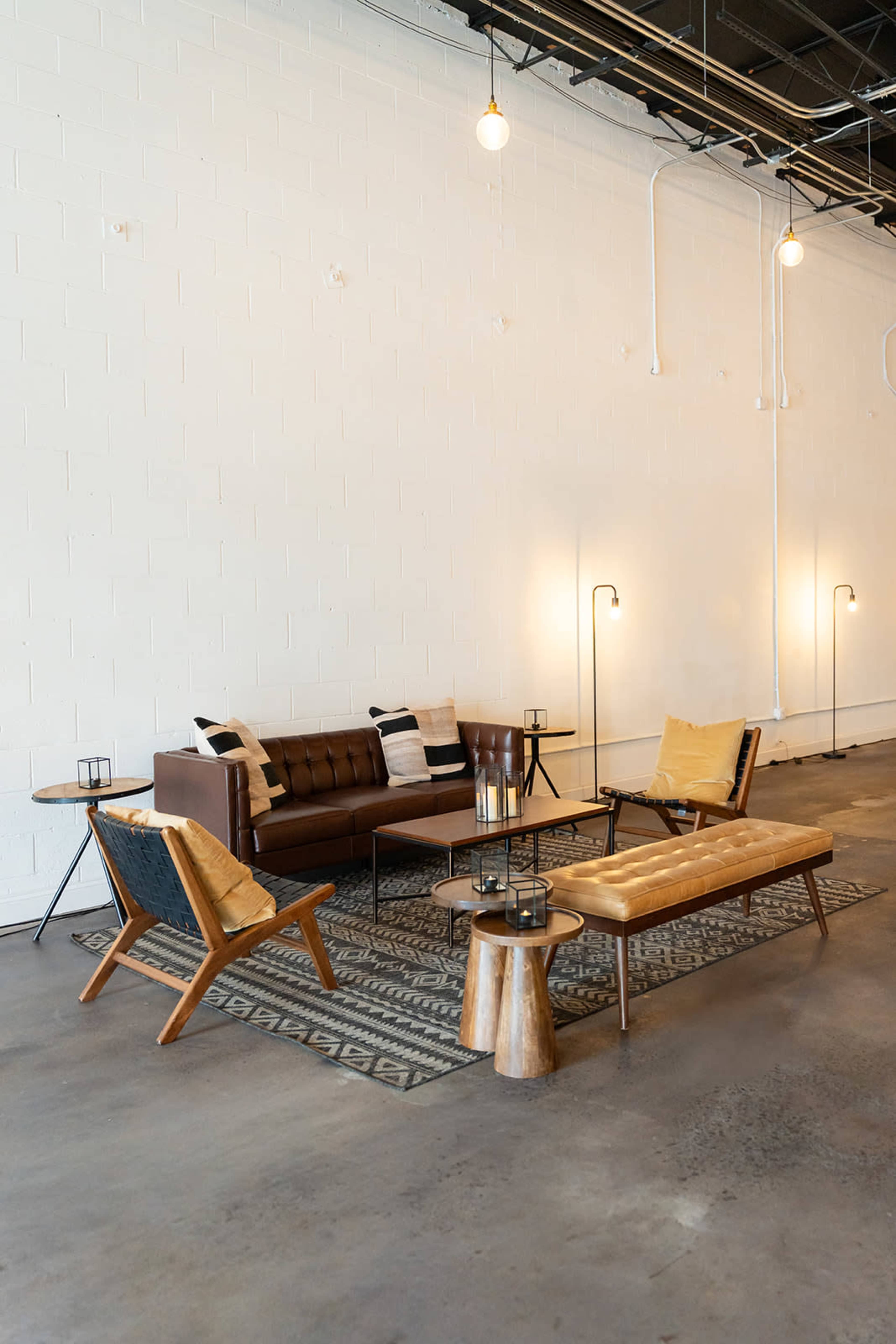 The image shows a modern, minimalist seating area with a brown leather sofa, wooden benches, and metal accents arranged on a concrete floor in an industrial-style space.