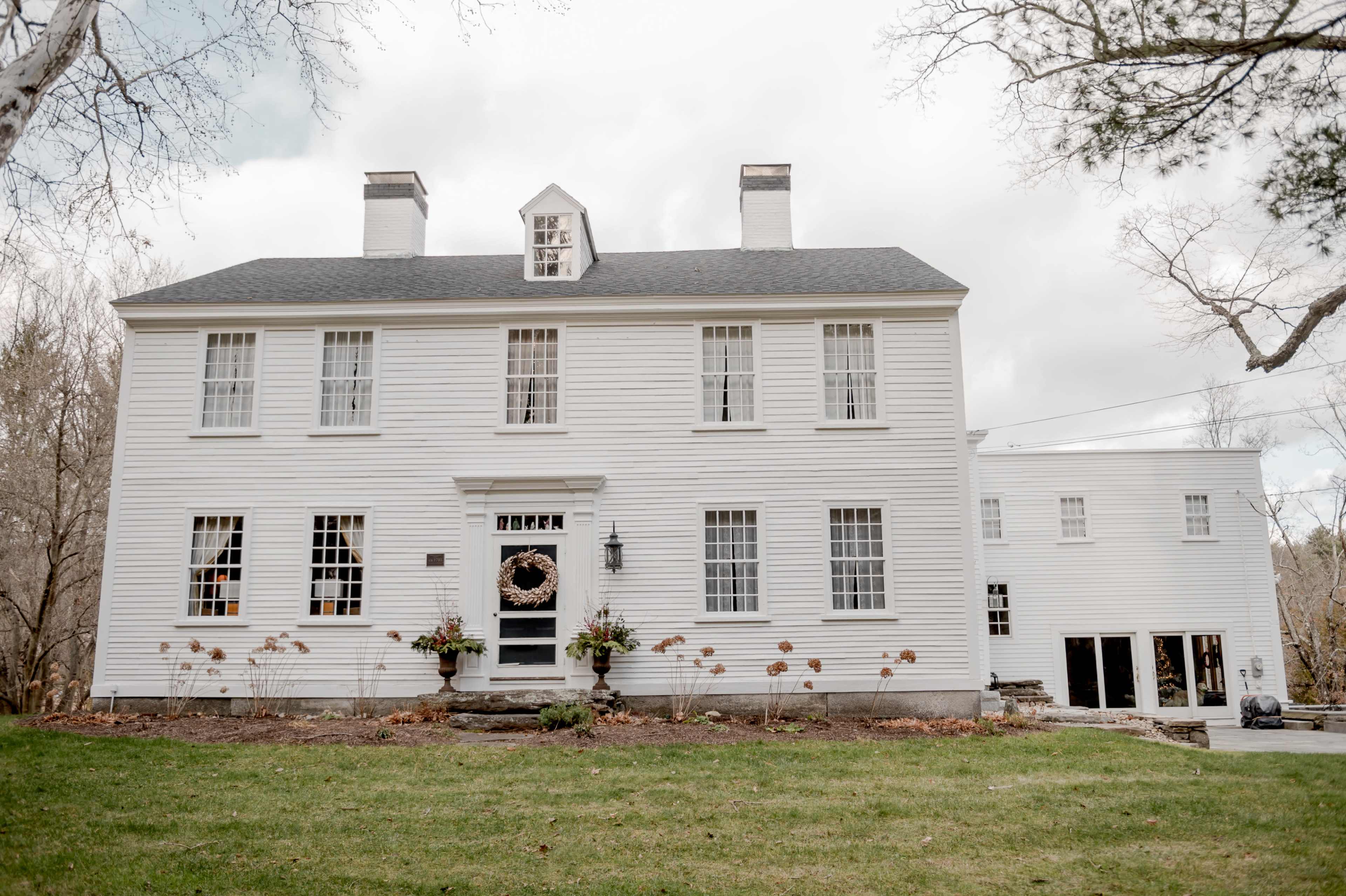 A large white colonial-style house with multiple windows and a front door adorned with a wreath, set on a green lawn under a cloudy sky.