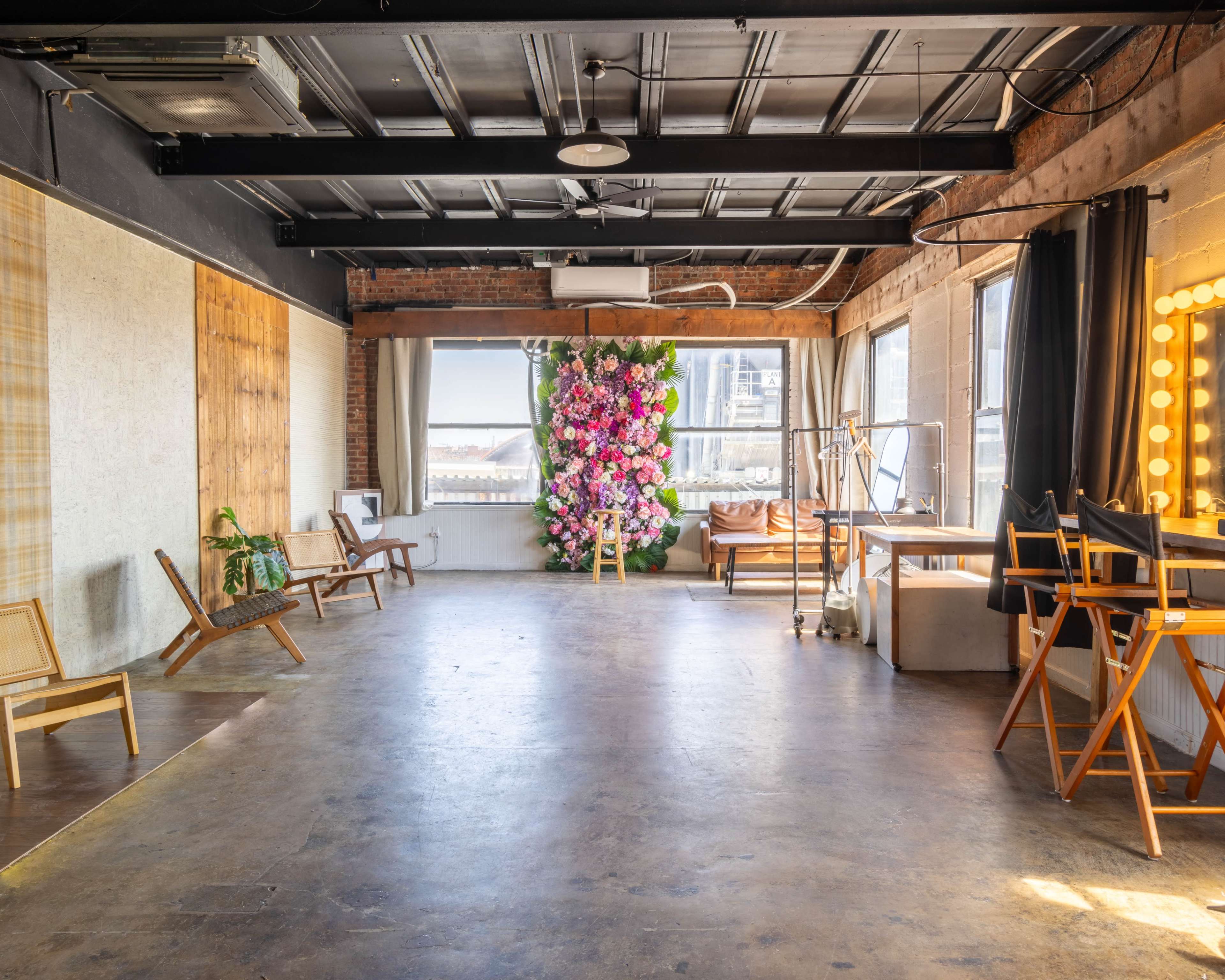 The image shows a spacious, well-lit room with wooden and metal furniture, featuring a floral backdrop and large windows.