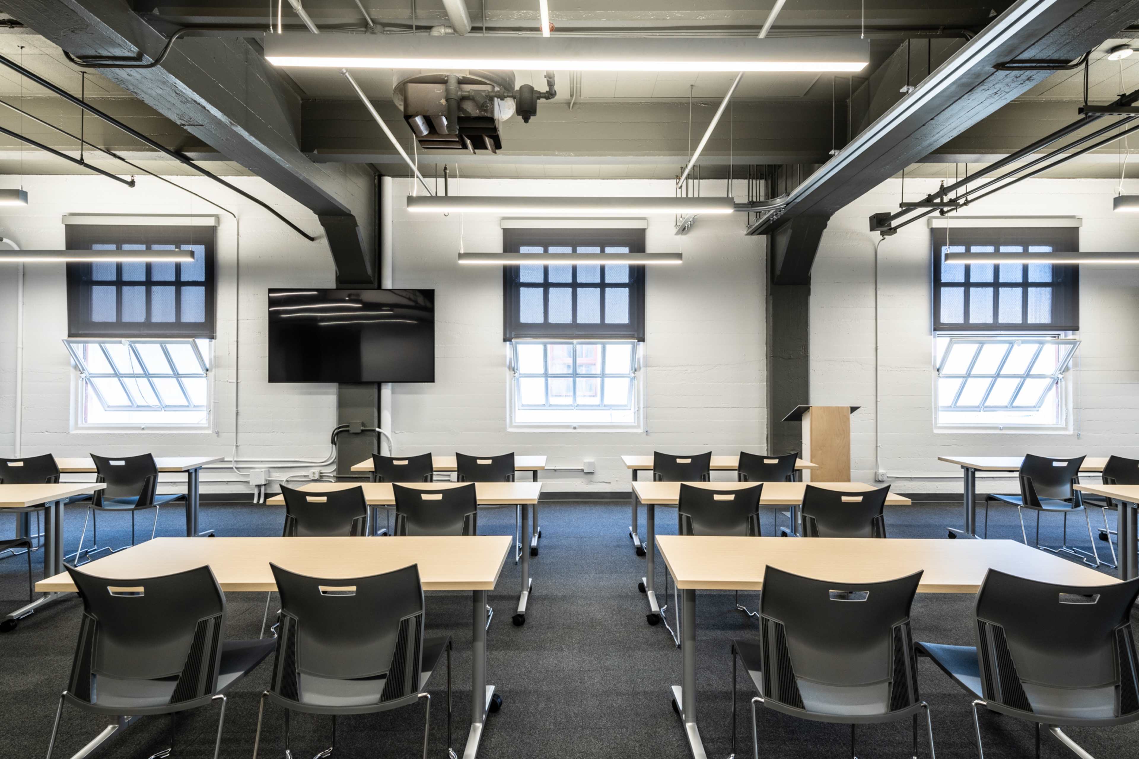 A modern classroom features rows of tables and chairs facing a presentation screen, with large windows allowing natural light.