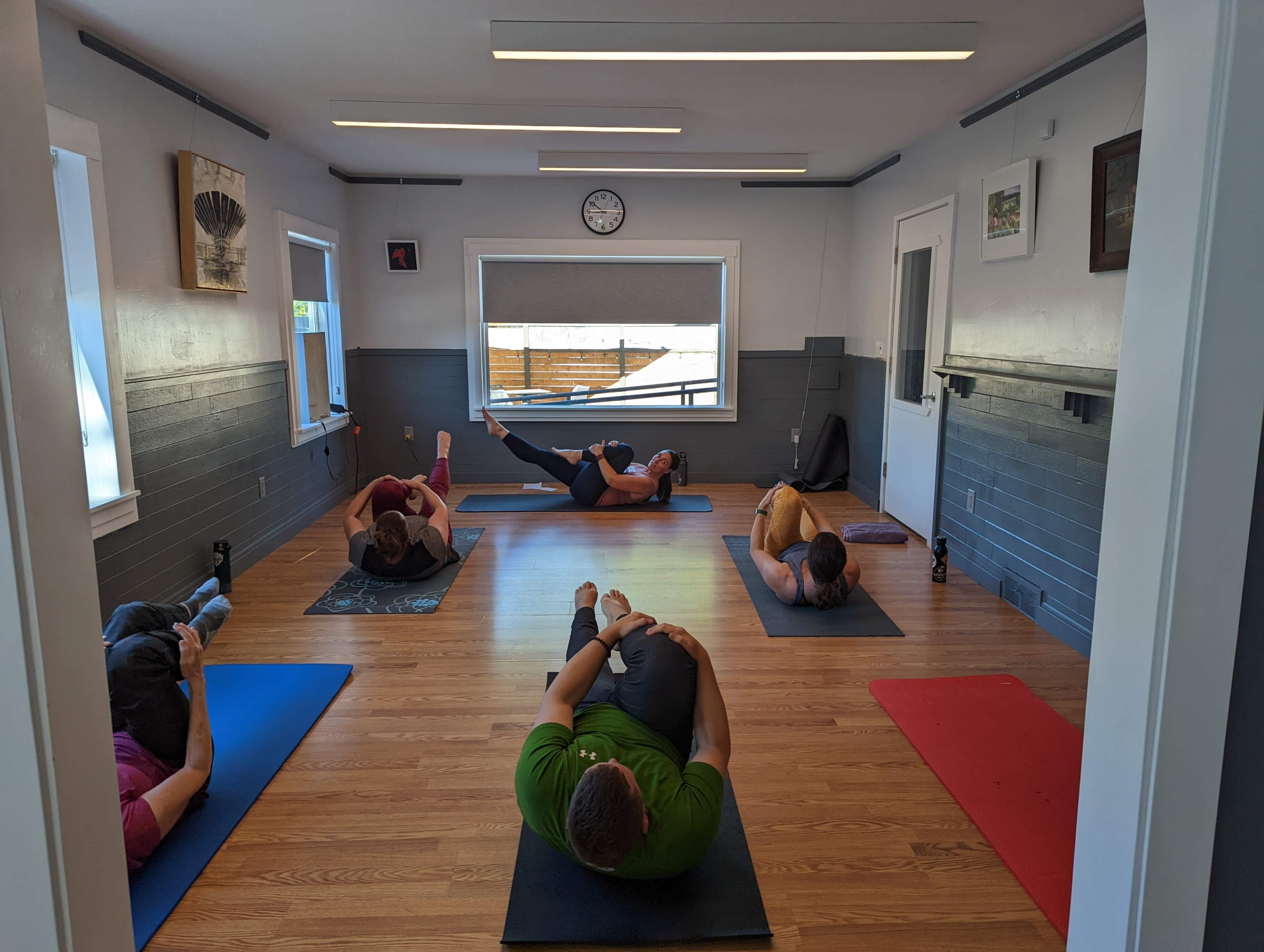 A group of six people is participating in a yoga or fitness class on mats in a well-lit room with wooden flooring and large windows.