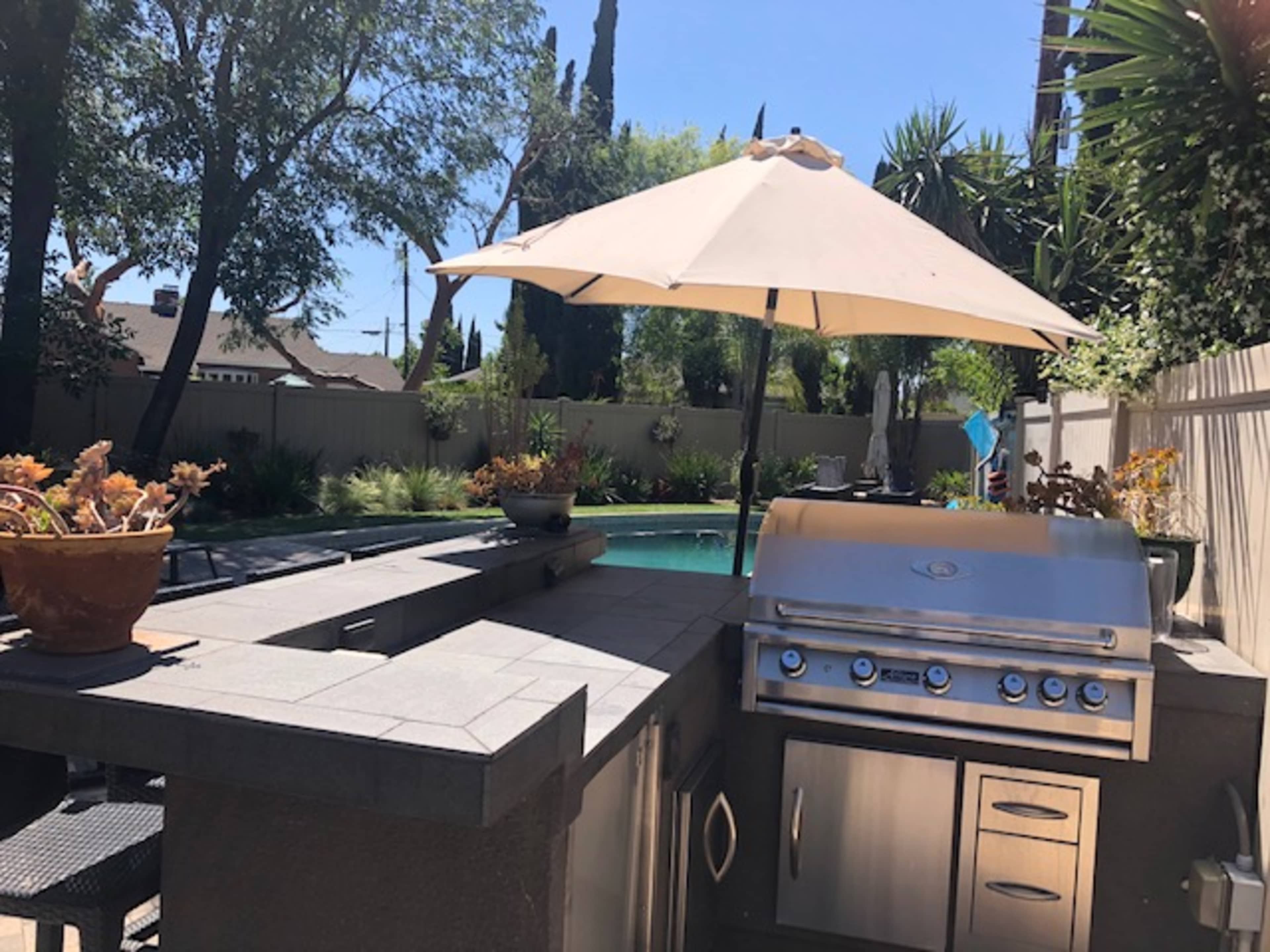 The image shows a backyard featuring a stainless steel barbecue grill under an umbrella, with a swimming pool and landscaped greenery in the background.