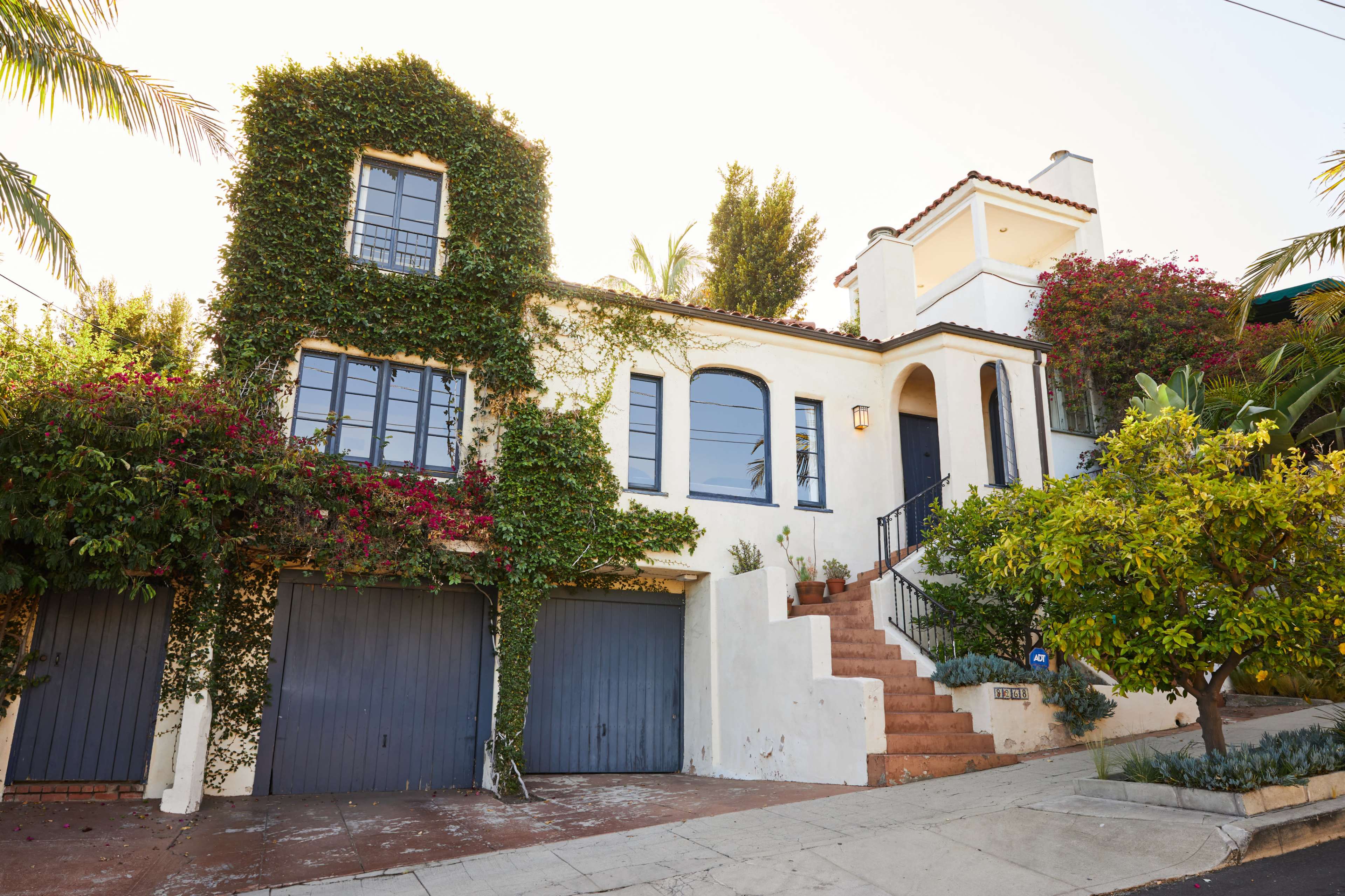 A two-story house adorned with climbing vines and colorful plants leads up to a set of stairs.