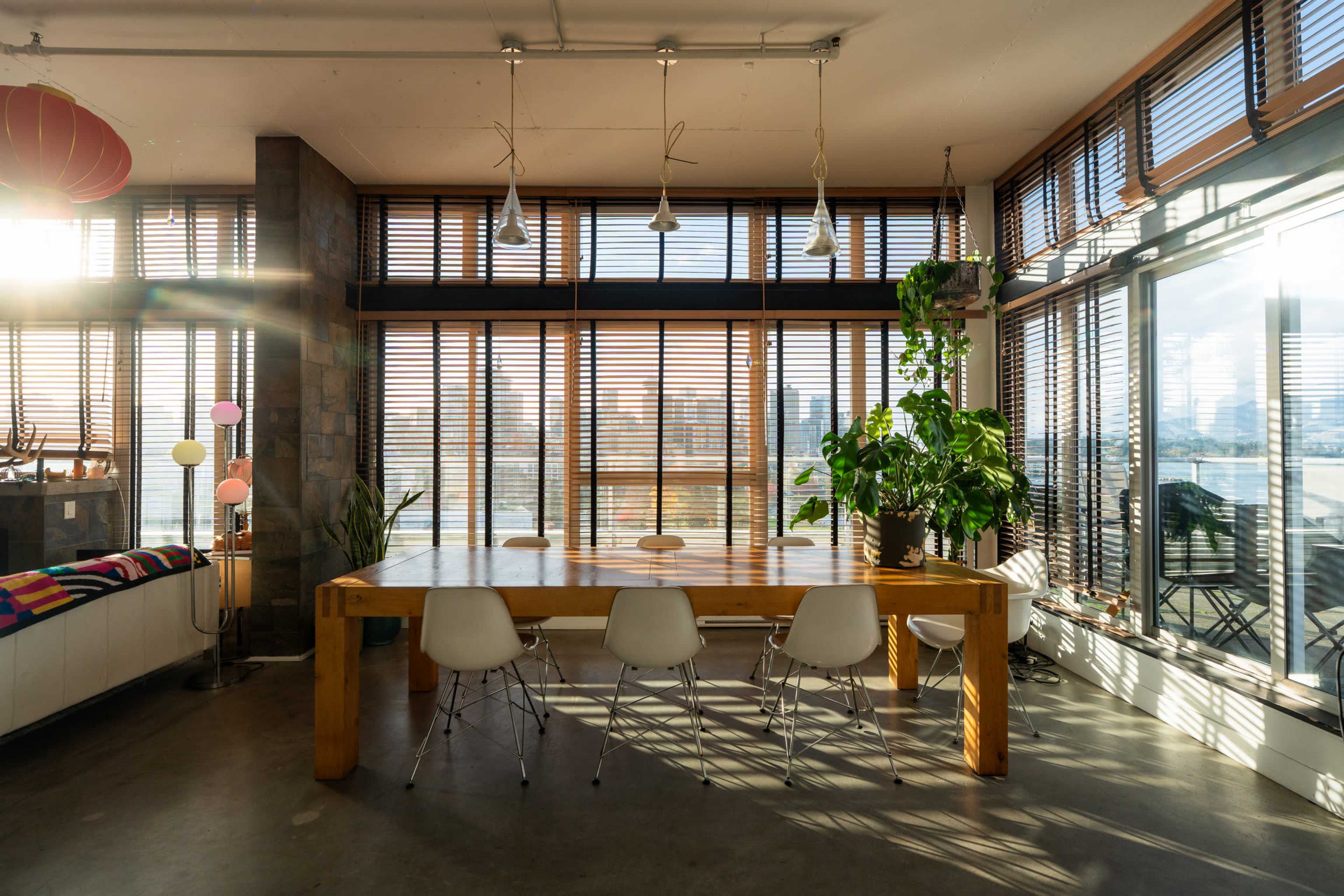 A large wooden dining table surrounded by white chairs is illuminated by sunlight streaming through tall windows with wooden blinds.
