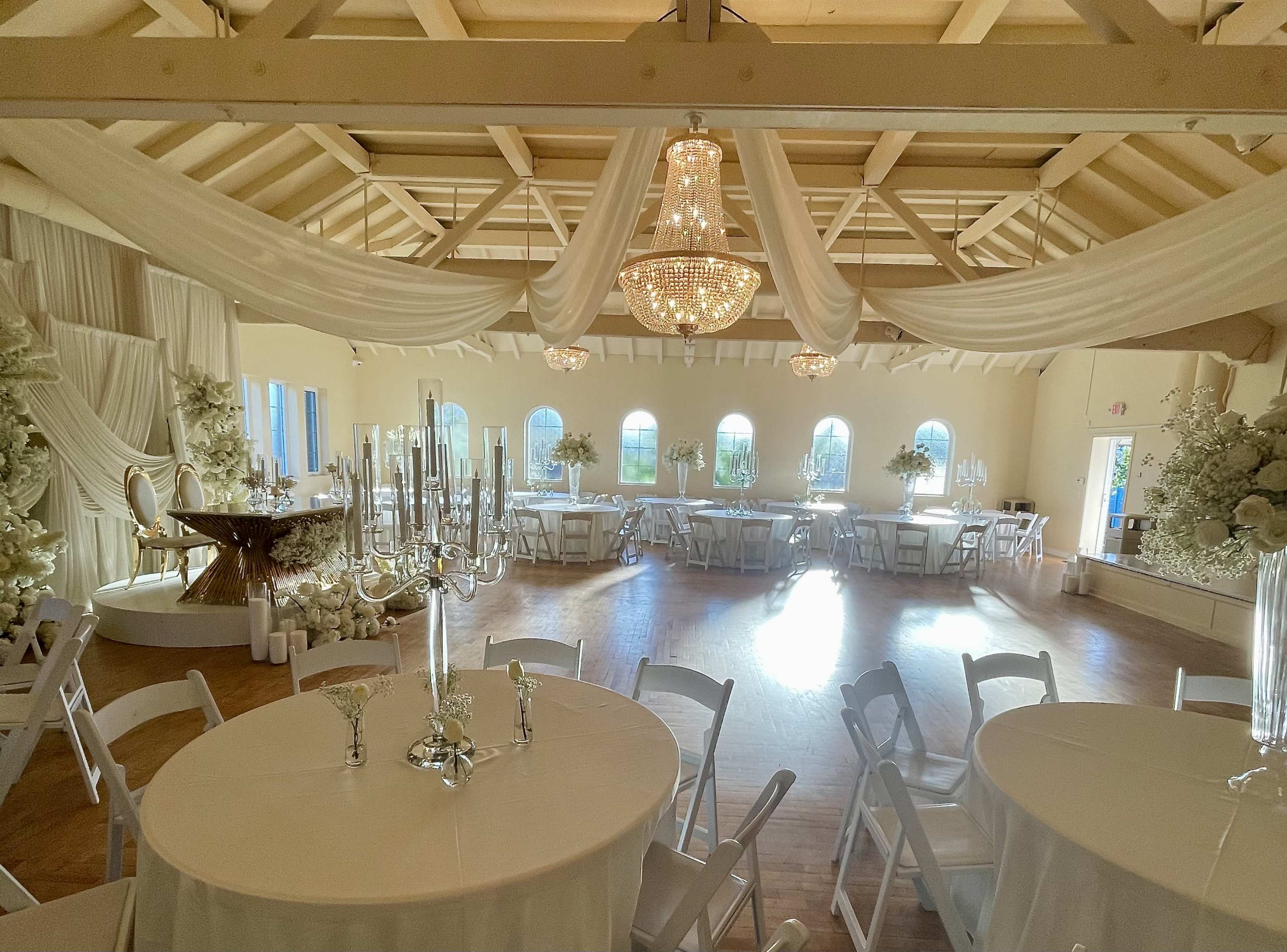 The image shows a spacious, elegantly decorated event hall with white drapery, chandeliers, and round tables set for a formal gathering.