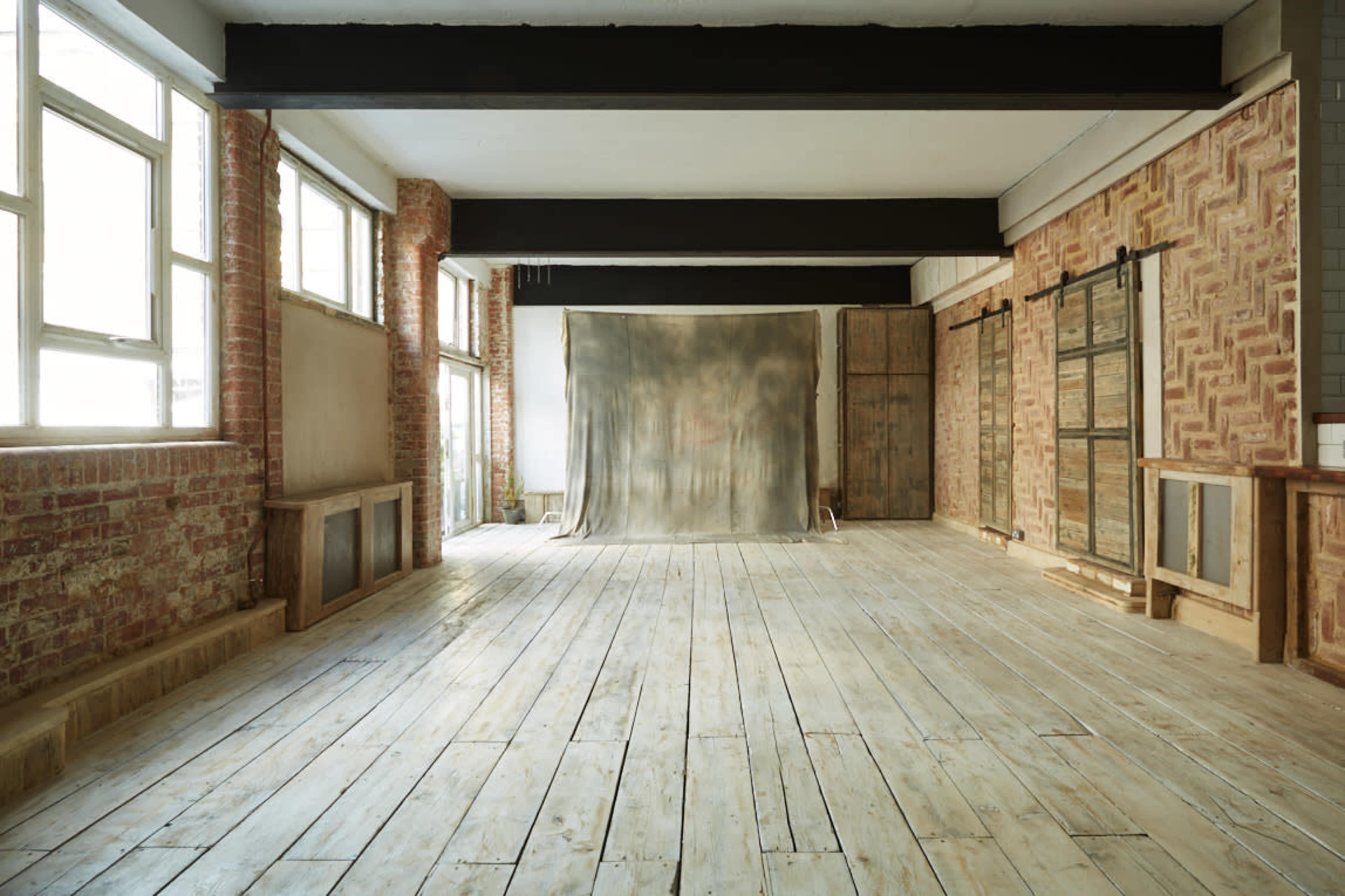An empty studio space with large windows, exposed brick walls, and a wooden floor, featuring a plain backdrop in the center.