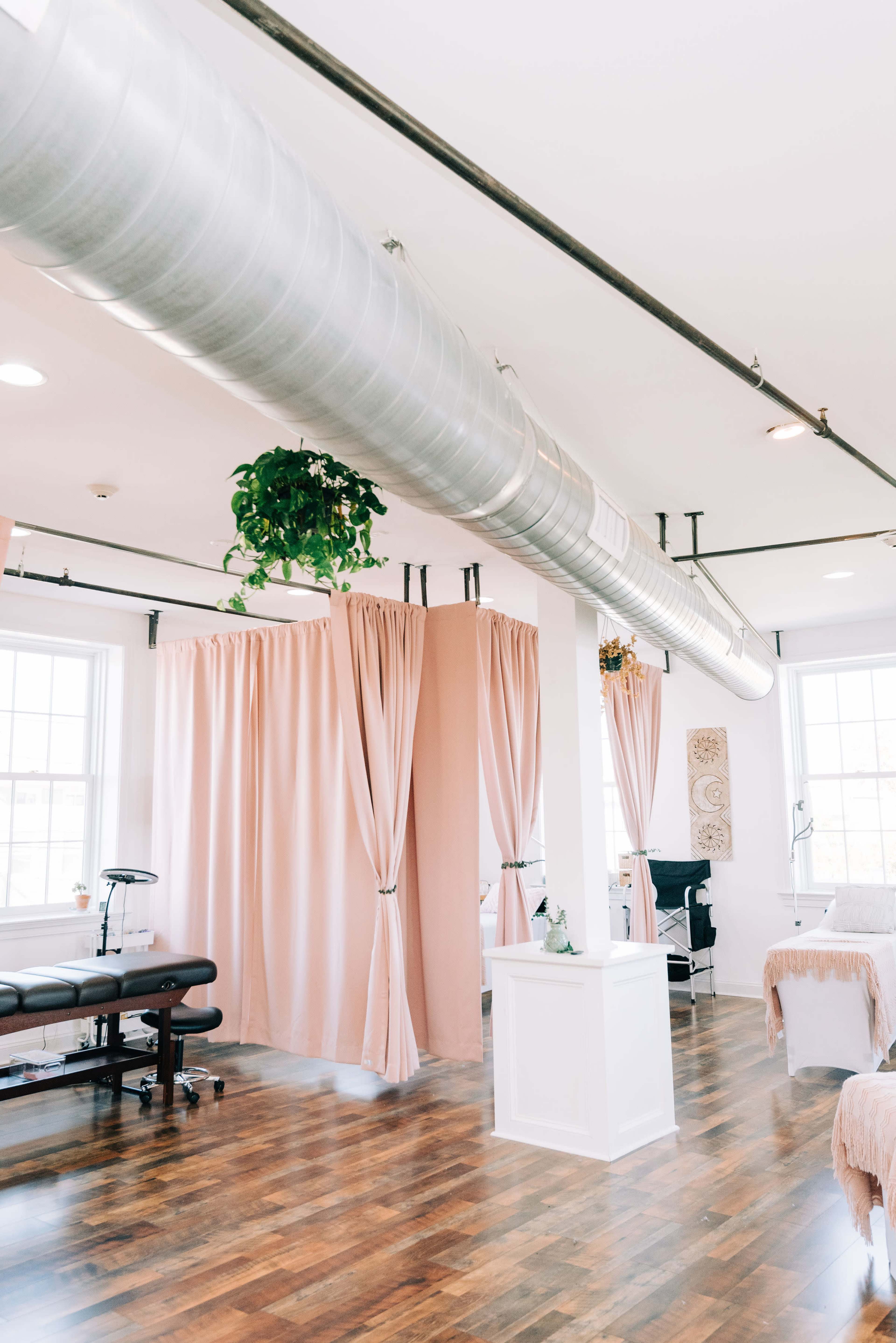 The image shows a modern indoor spa or treatment room with large windows, wooden flooring, and divided spaces by soft pink curtains.