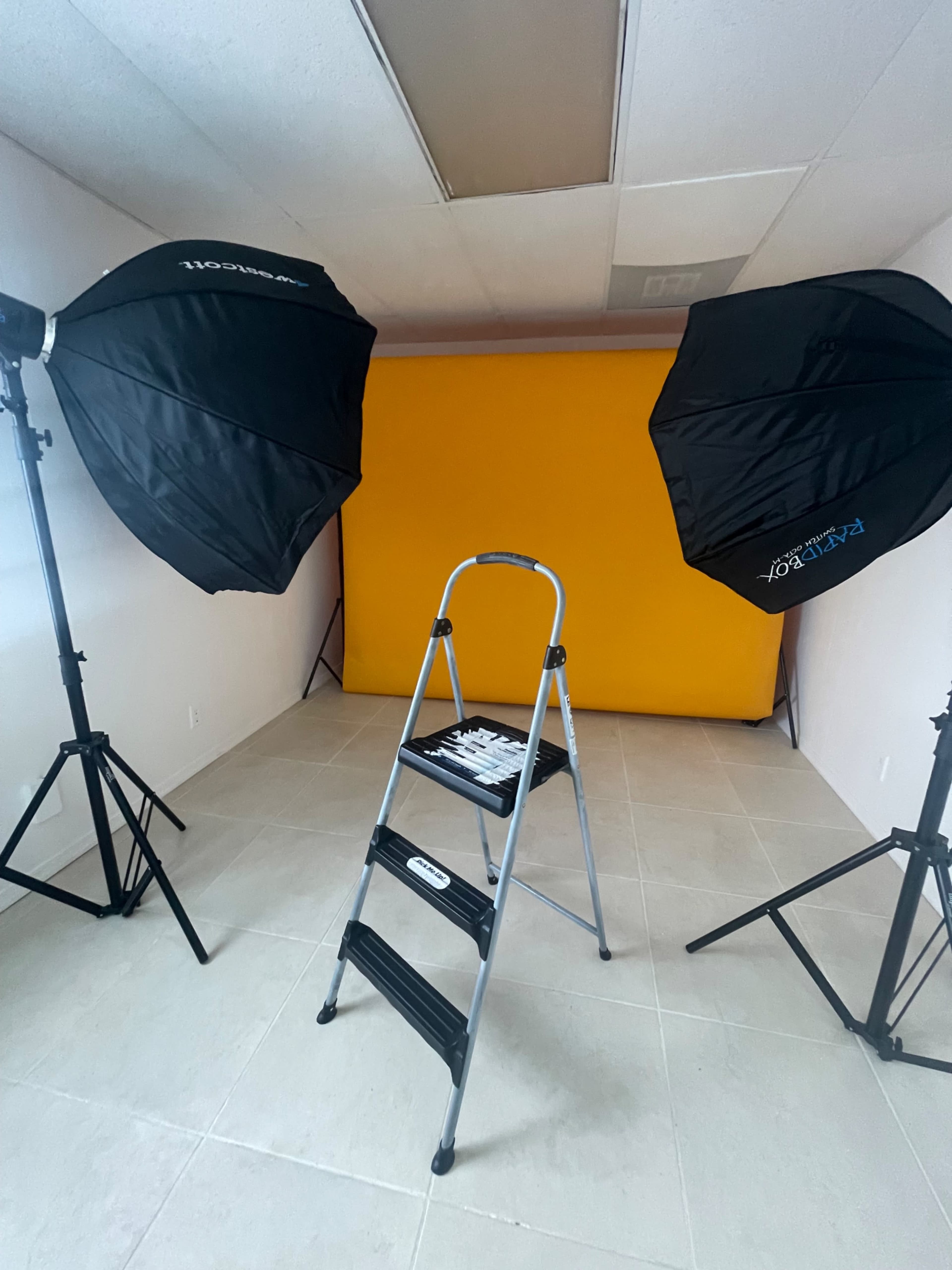 A photo studio setup features a yellow backdrop, two light sources on stands, and a step ladder in the center.