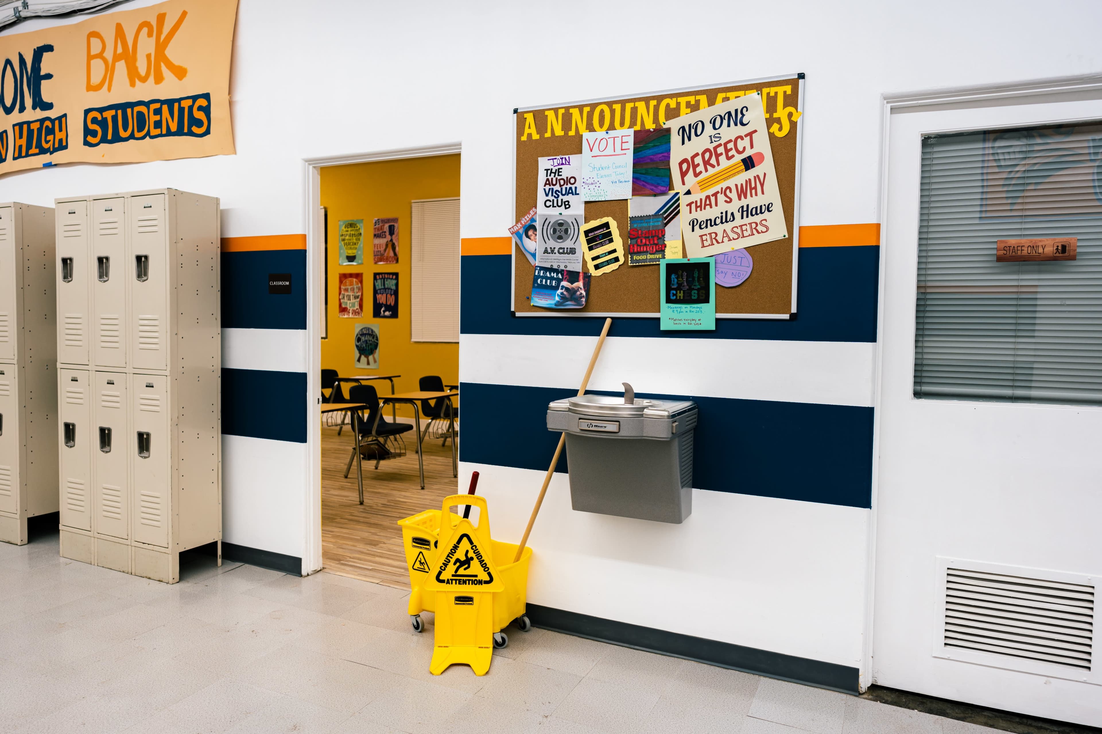 A school hallway features lockers, an announcements board with posters, a trash can, and a caution sign.