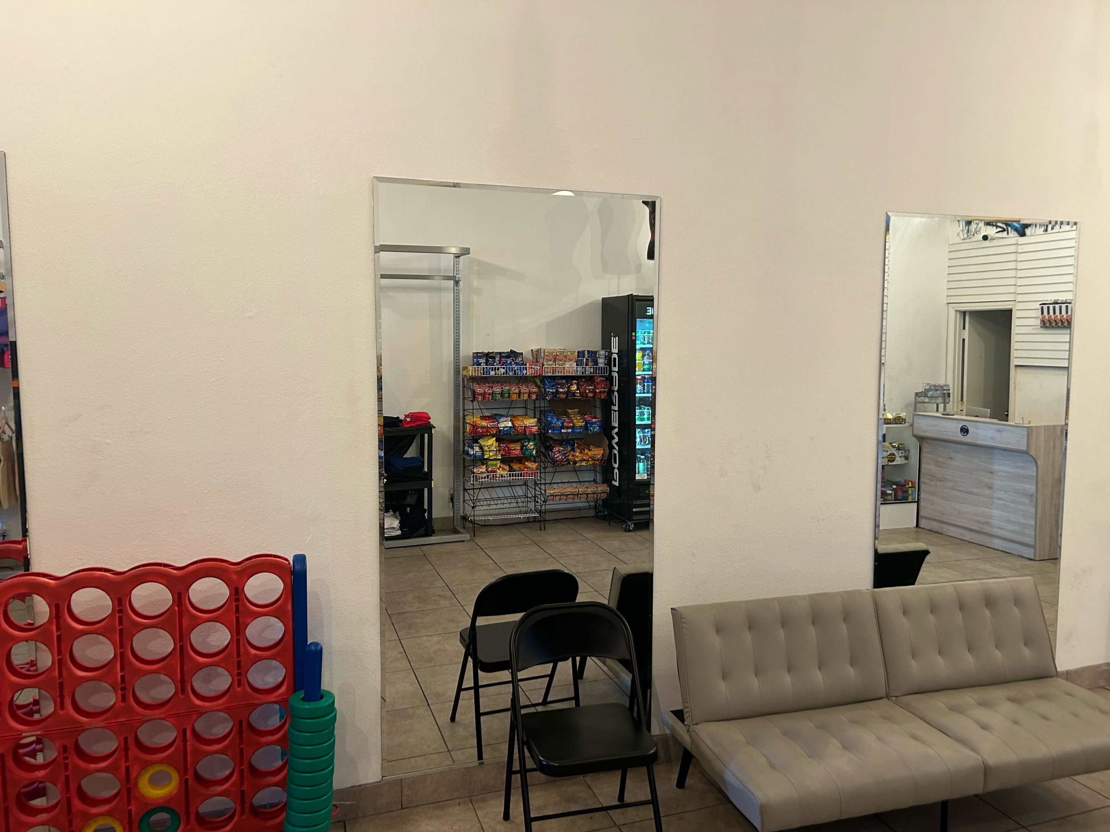 The image shows a casual waiting area with a gray sofa, a large Connect Four game, and a view of vending machines and shelves filled with snacks through two mirrors on the wall.