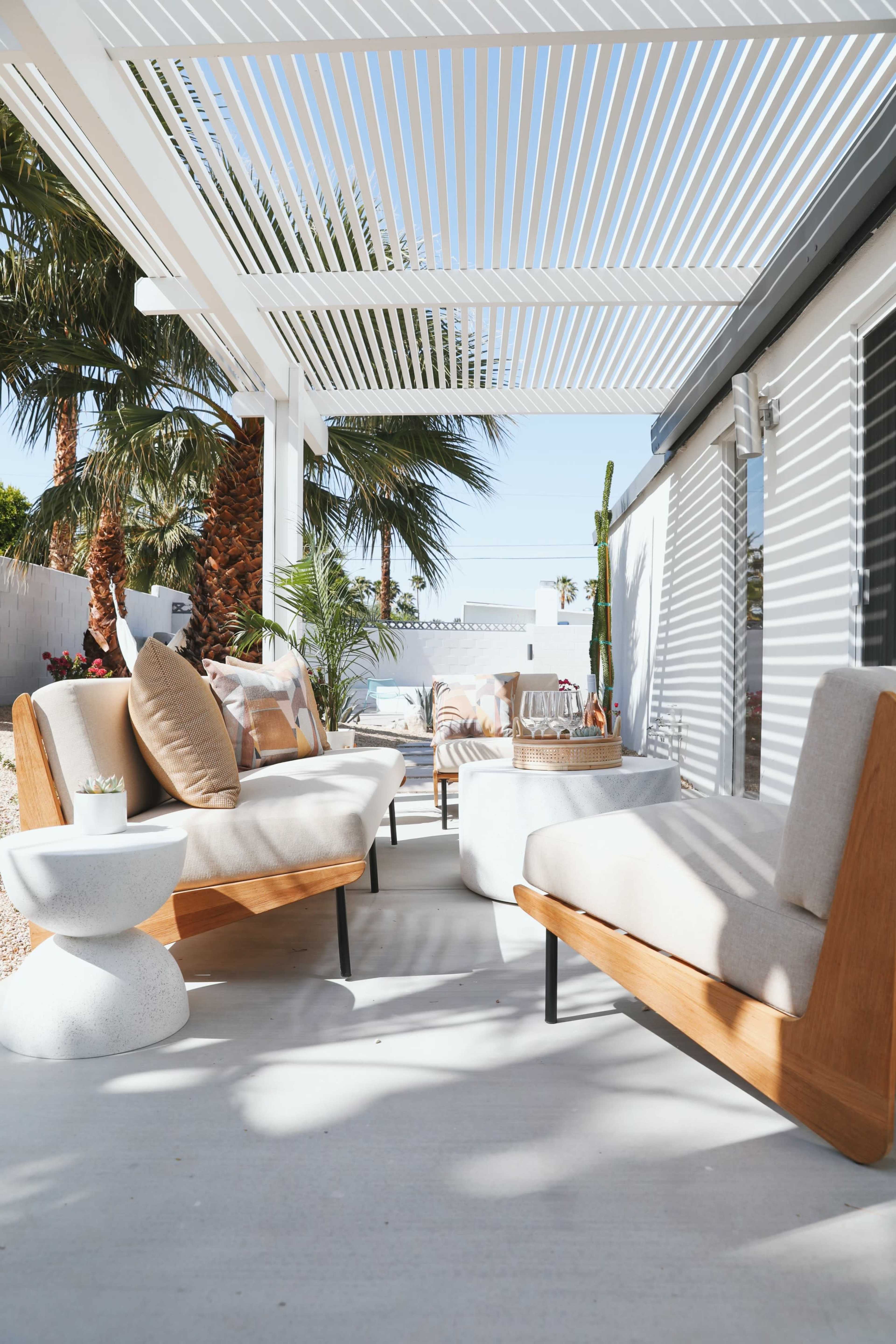 A modern patio features wooden seating, a round table, and decorative plants under a slatted pergola with sunlight streaming through.