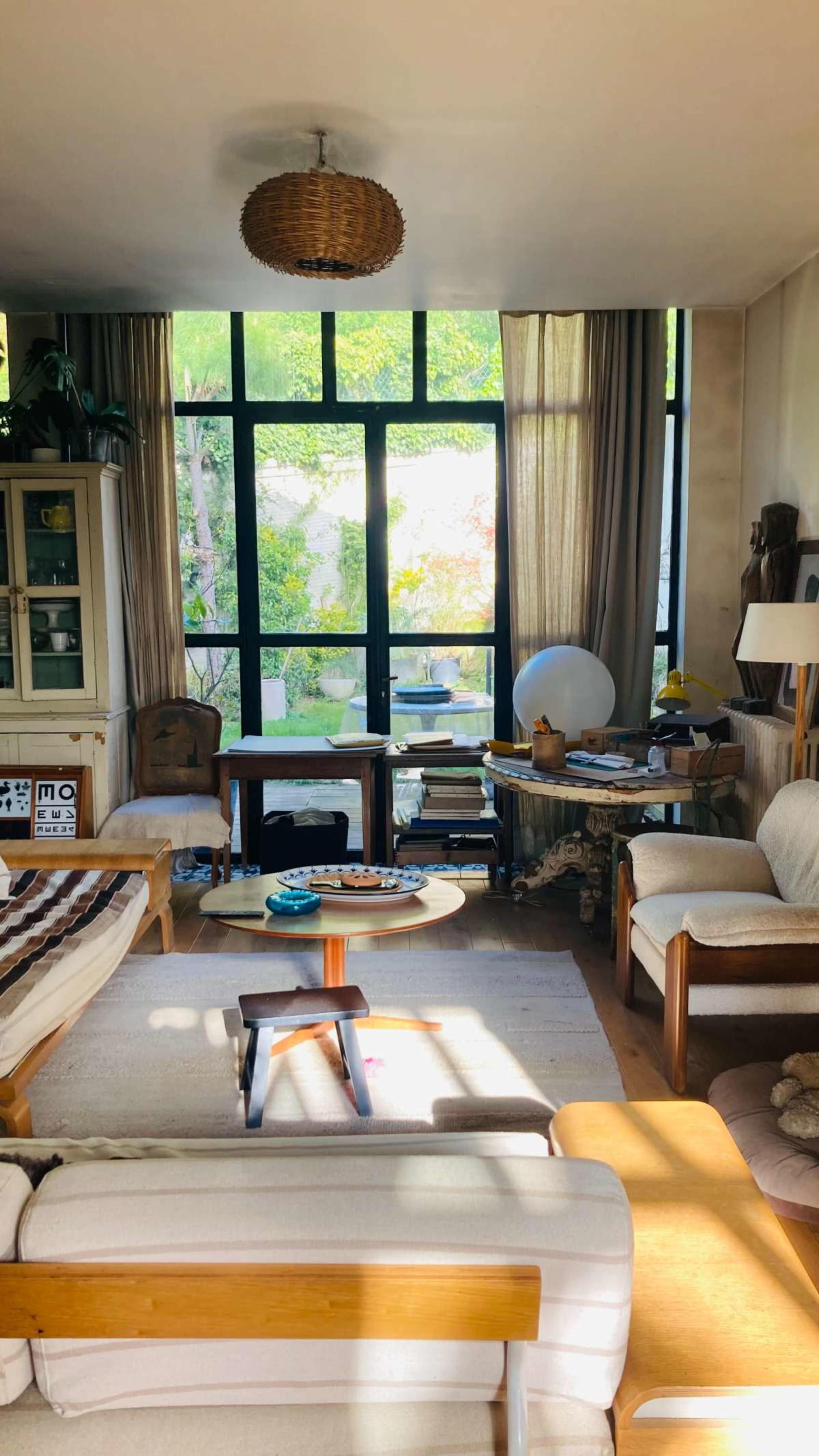 A cozy living room features large windows, light-colored furniture, and various decorative items, with sunlight streaming in from outside.