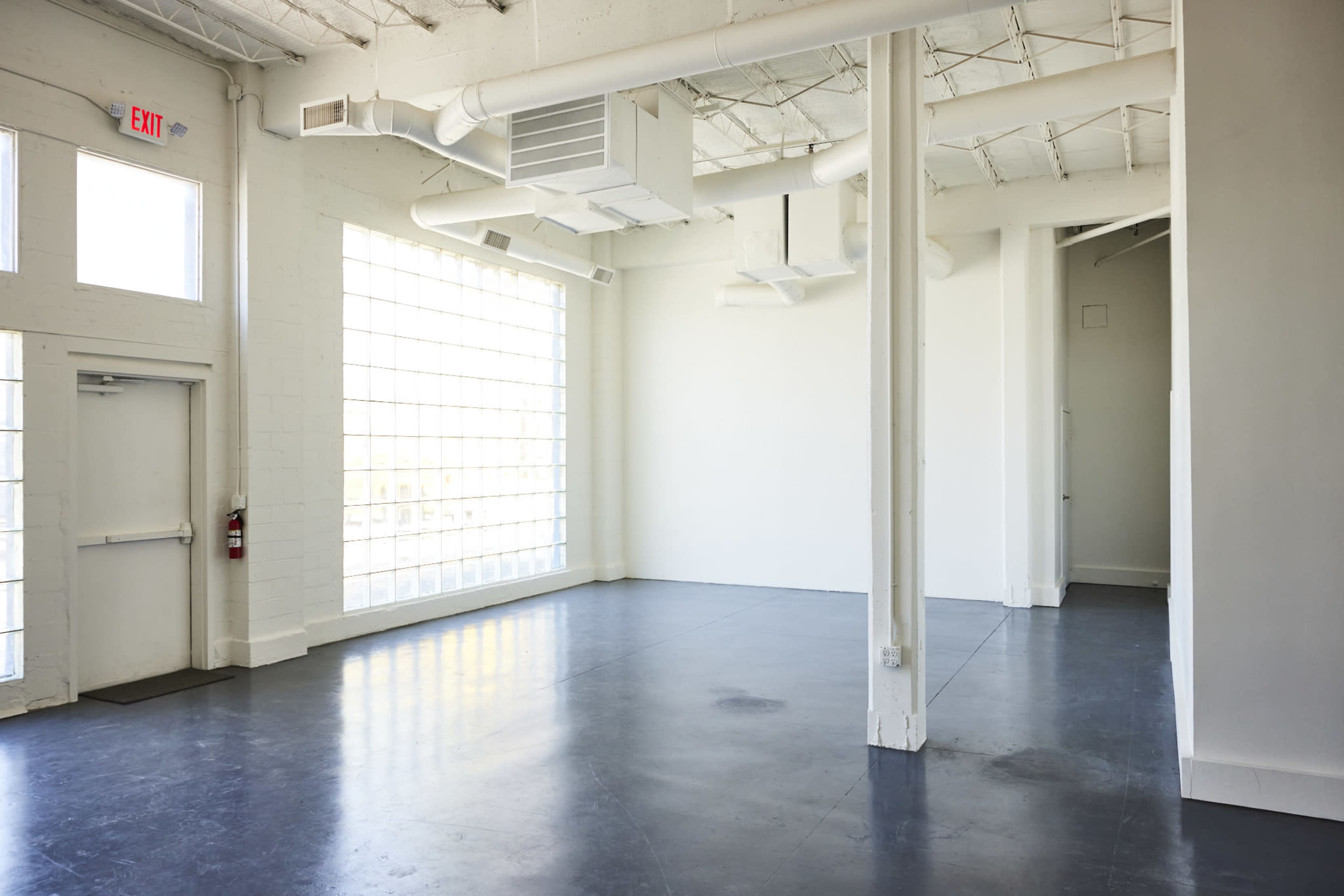 The image shows a spacious, empty interior with a polished black floor, large windows letting in natural light, and a simple layout featuring exposed ductwork and white walls.