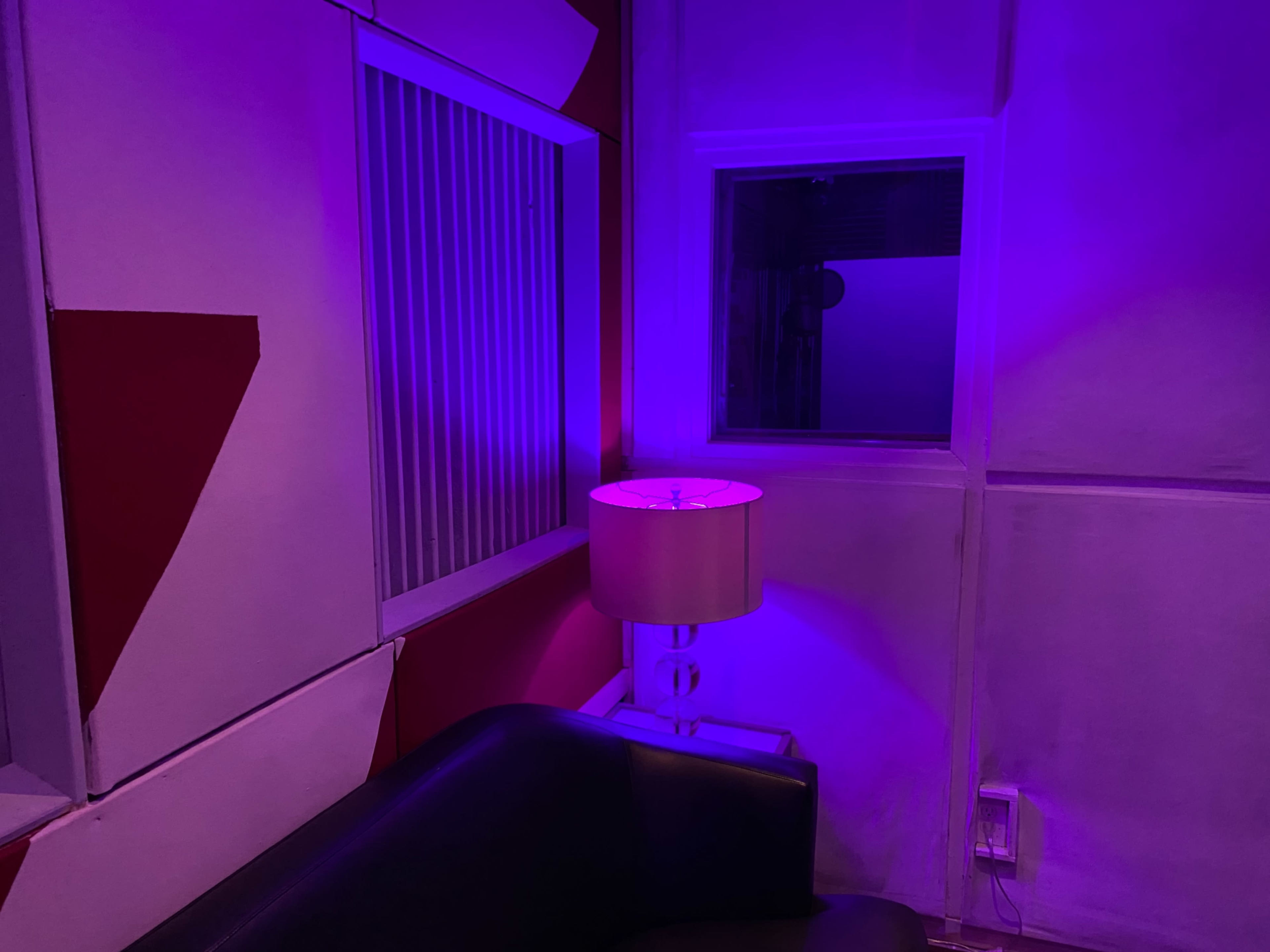 The image shows a corner of a room with a pink lamp illuminating a section of wall painted in red and white, alongside a window and a black couch.