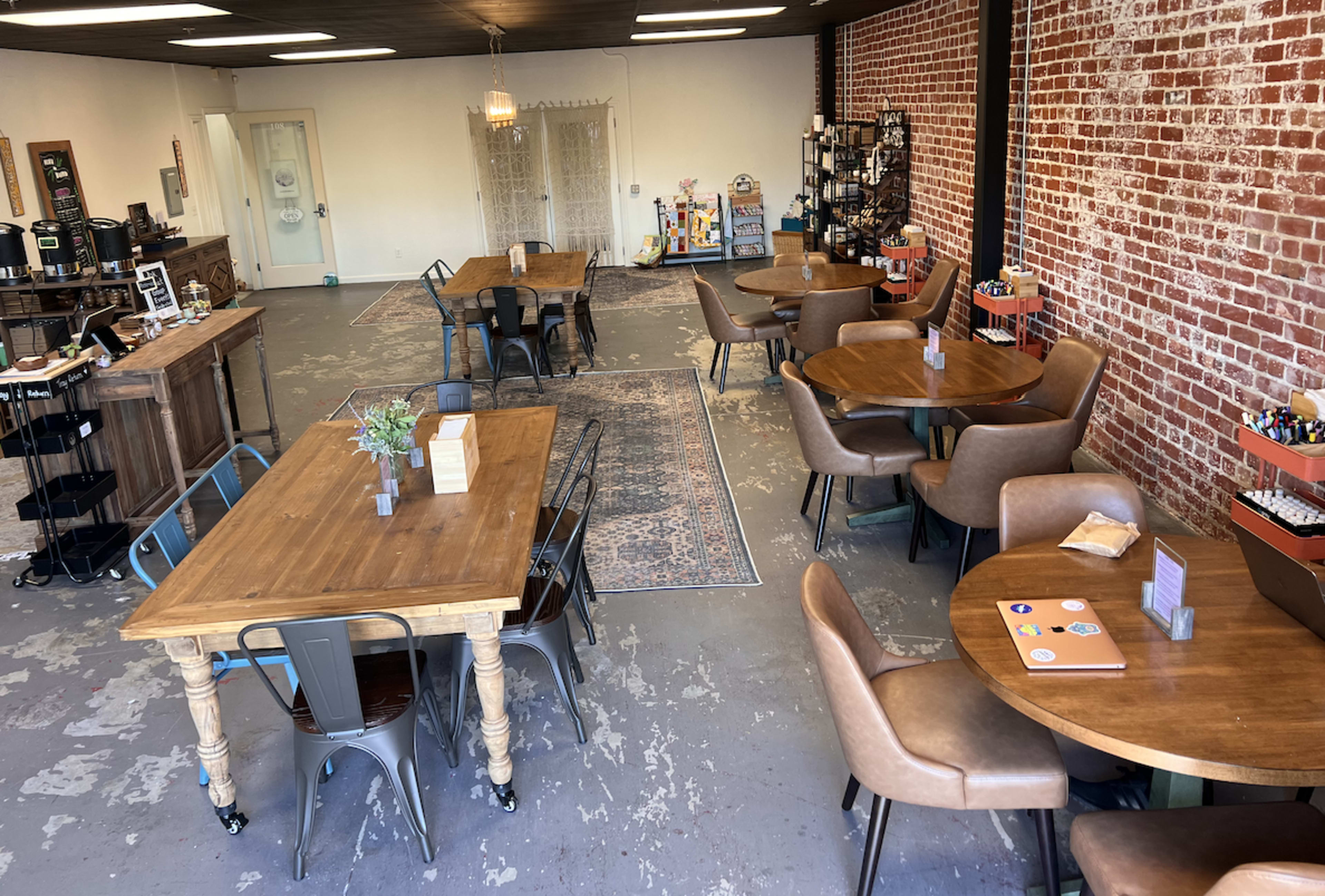 A spacious interior of a cafe or community space with wooden tables, chairs, and a brick wall, featuring a counter and decorative items.