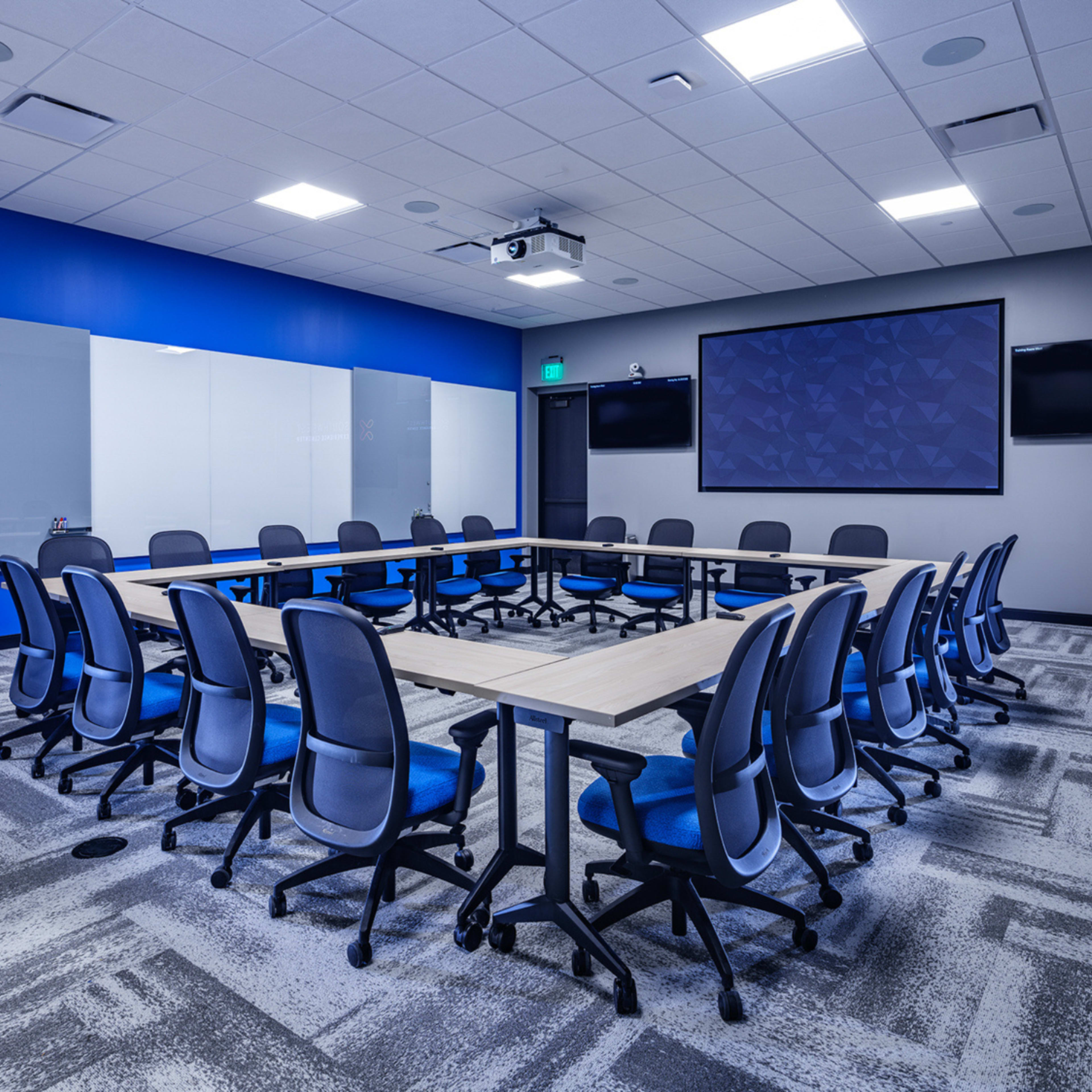 The image depicts a modern conference room featuring a rectangular arrangement of tables surrounded by ergonomic chairs, with a projector and screens mounted on the walls.